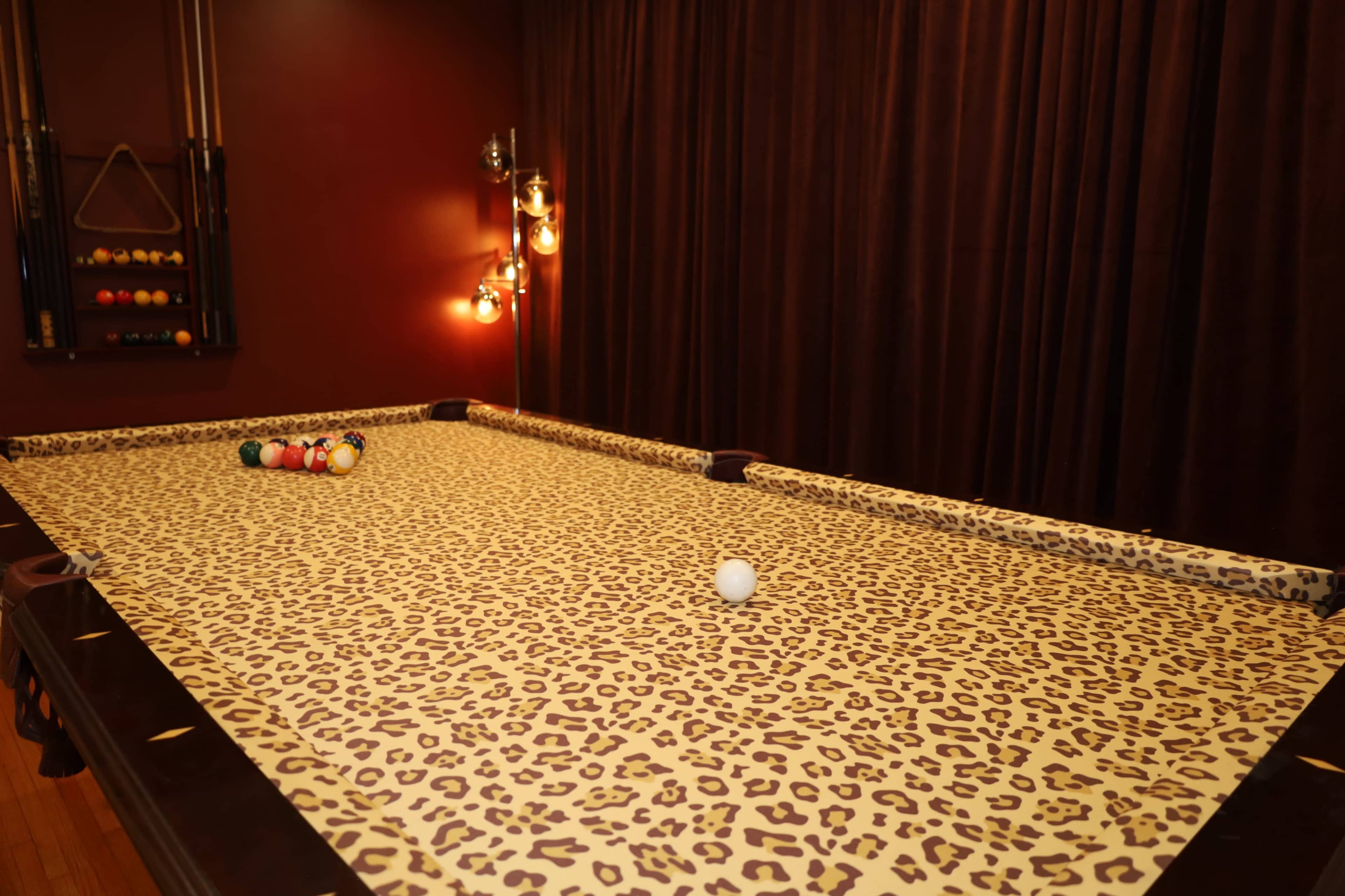 A billiard table with a leopard-print cover is set against a dark, textured wall, and a few colorful balls are positioned on the table.