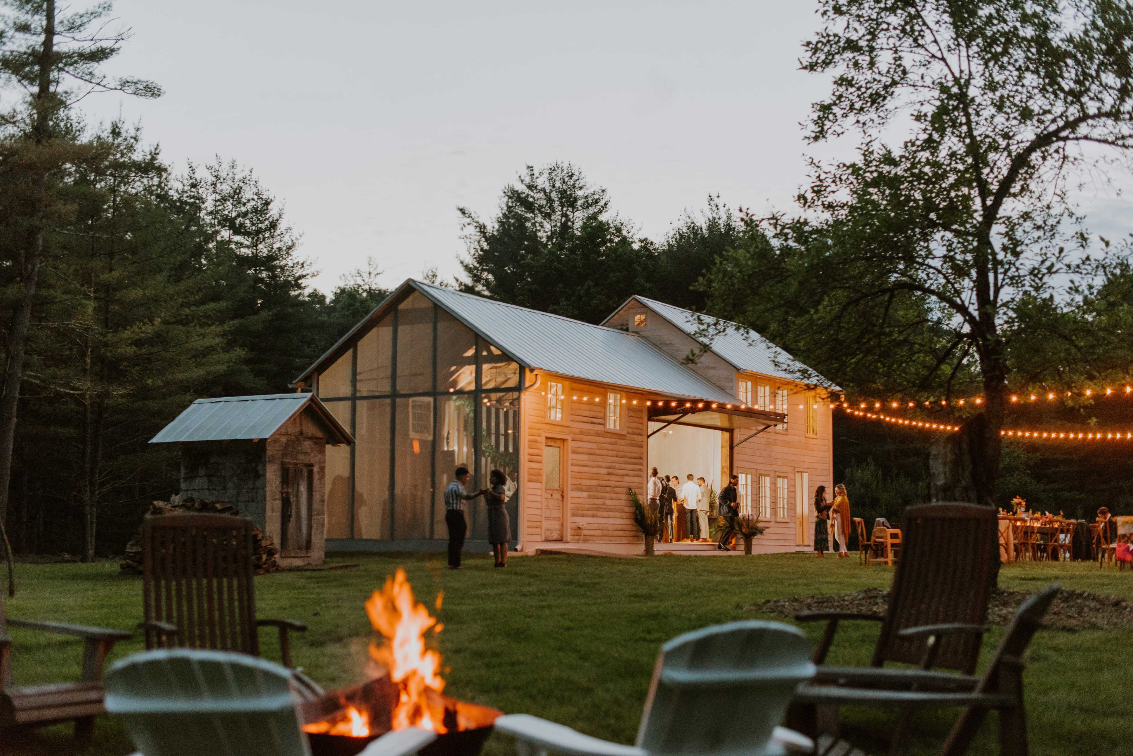 A wooden barn is illuminated in the evening with guests gathered outside around a fire pit and string lights overhead.