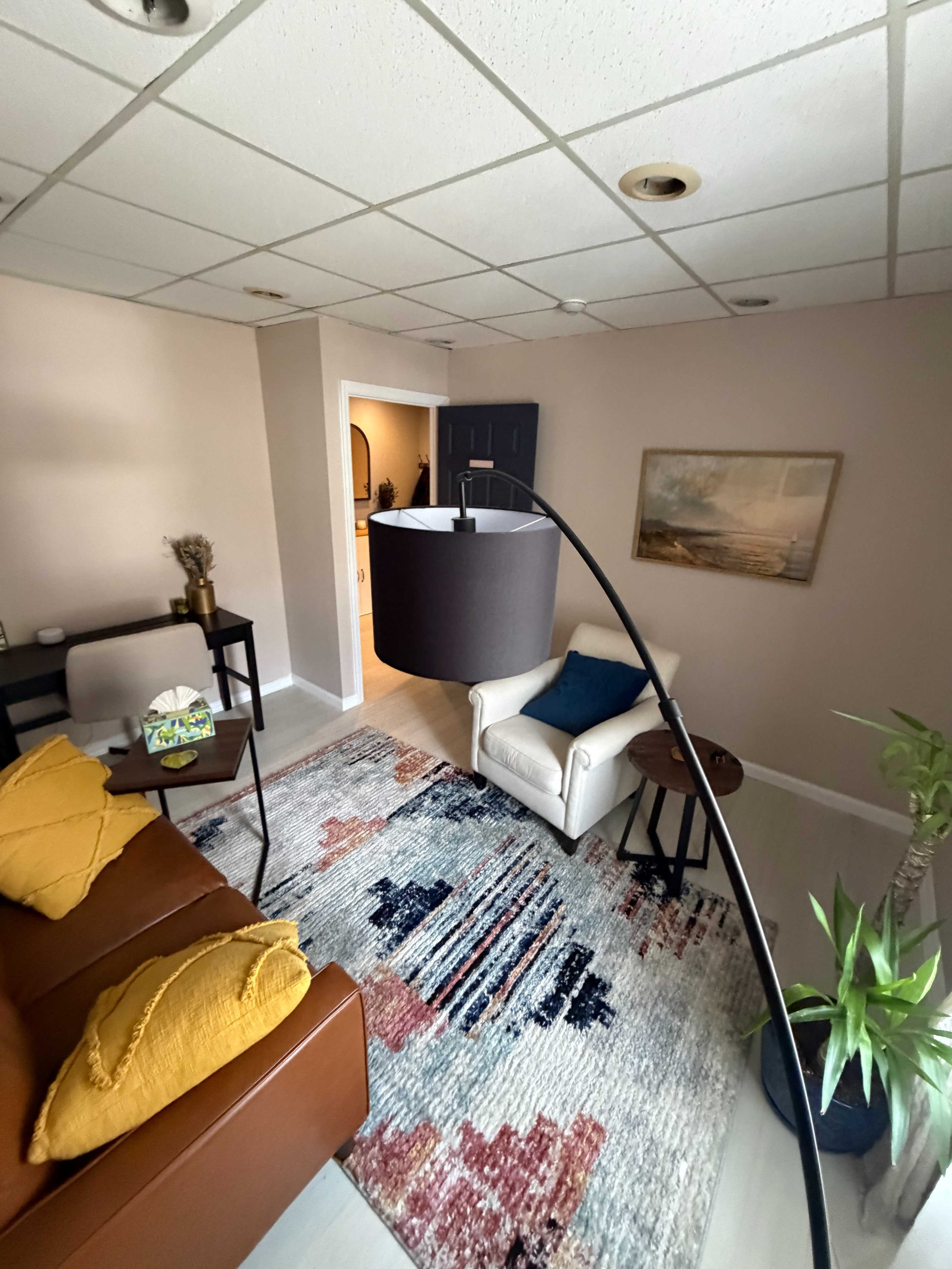 The image shows a living room with a brown sofa, a white armchair, a patterned rug, a floor lamp, and a view into a kitchen area through an open doorway.