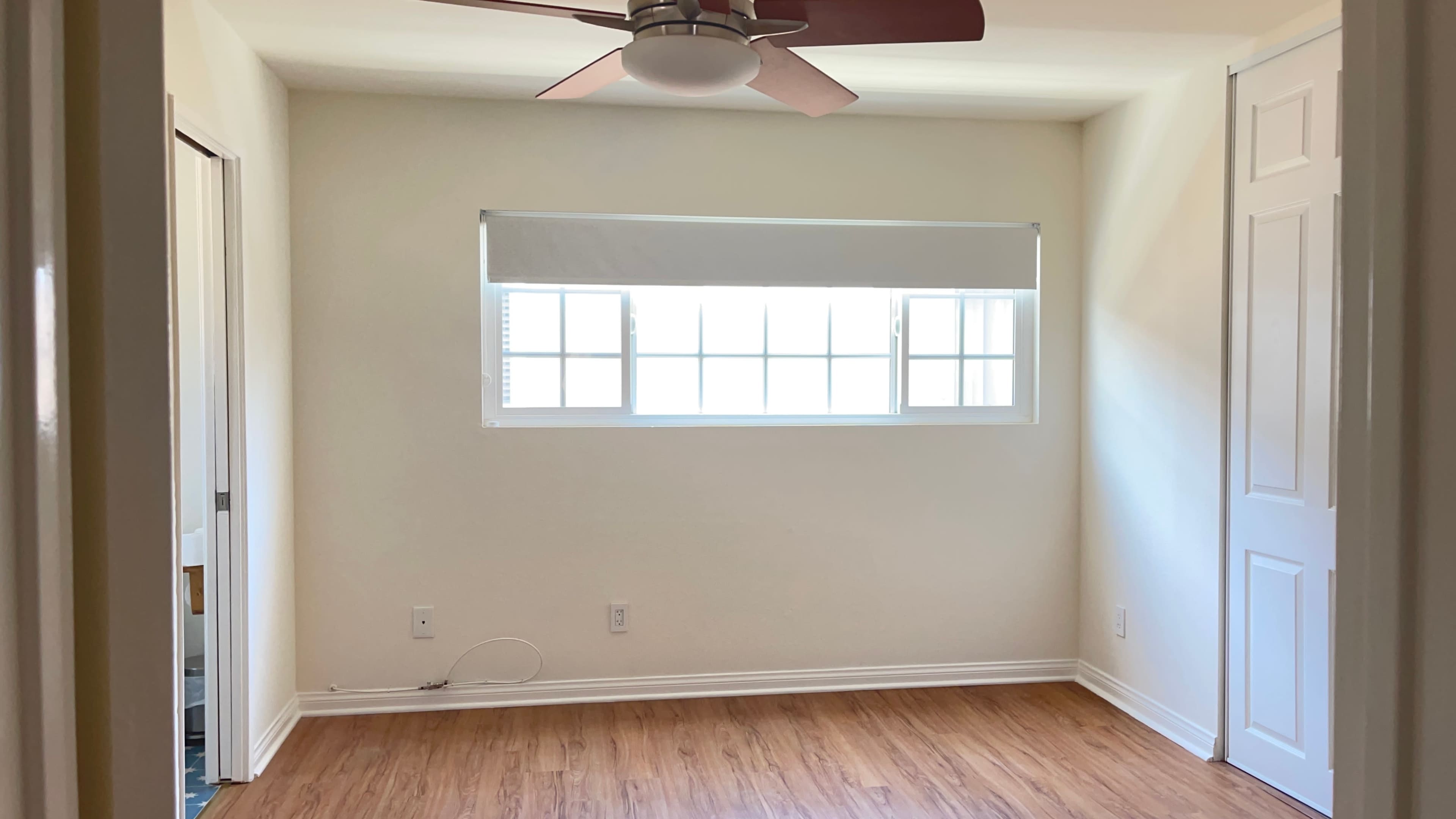 A bright room with a ceiling fan, a large window with blinds, and wooden flooring.
