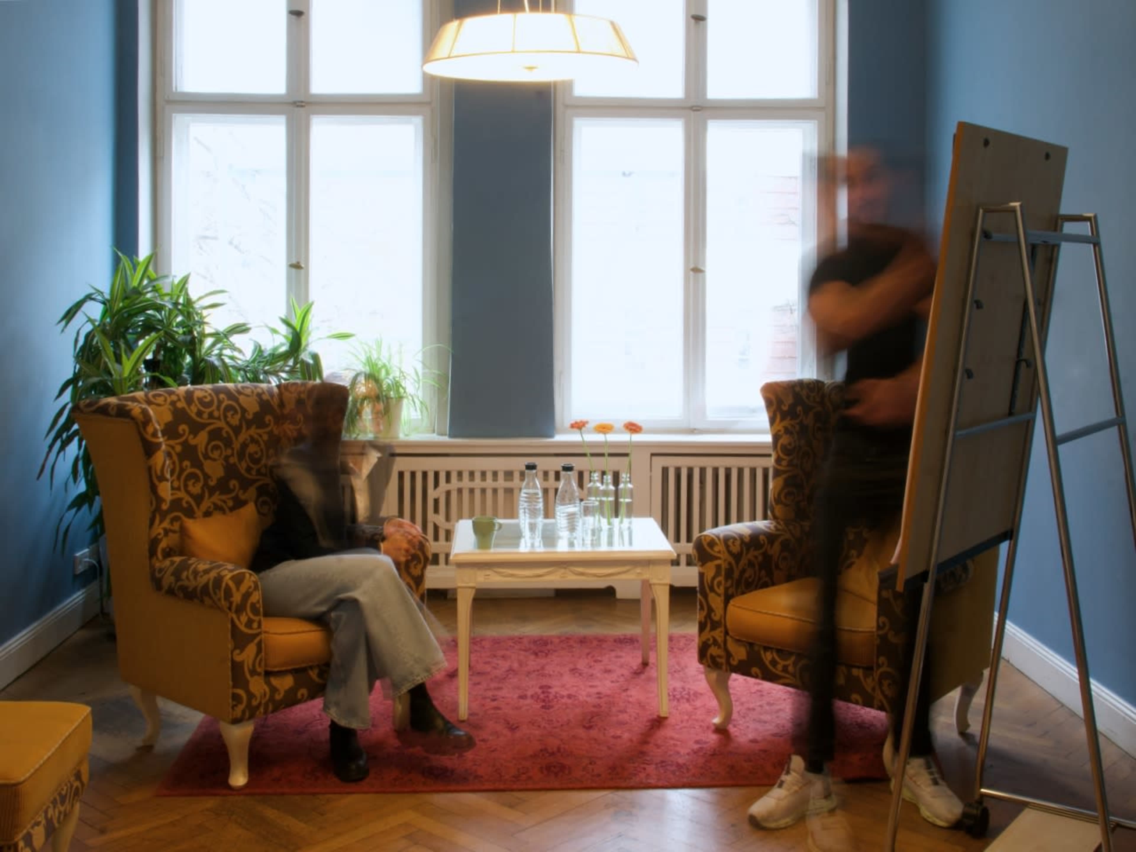 A person sits in a chair while another stands near a presentation board in a well-lit room with blue walls and a small coffee table.