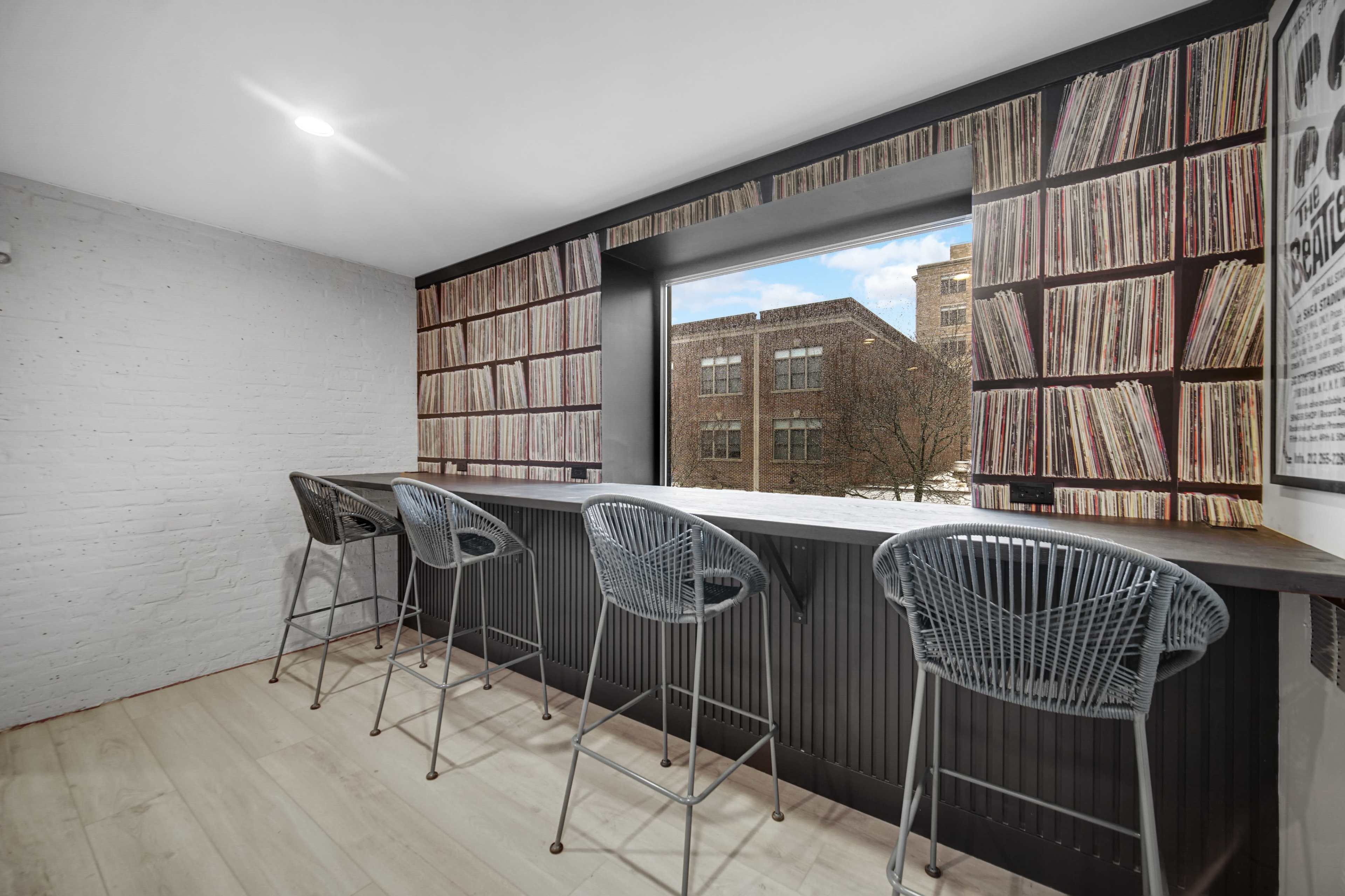 A modern bar area features four wire counter stools at a white countertop, next to a large window framed by a wall of vinyl records.