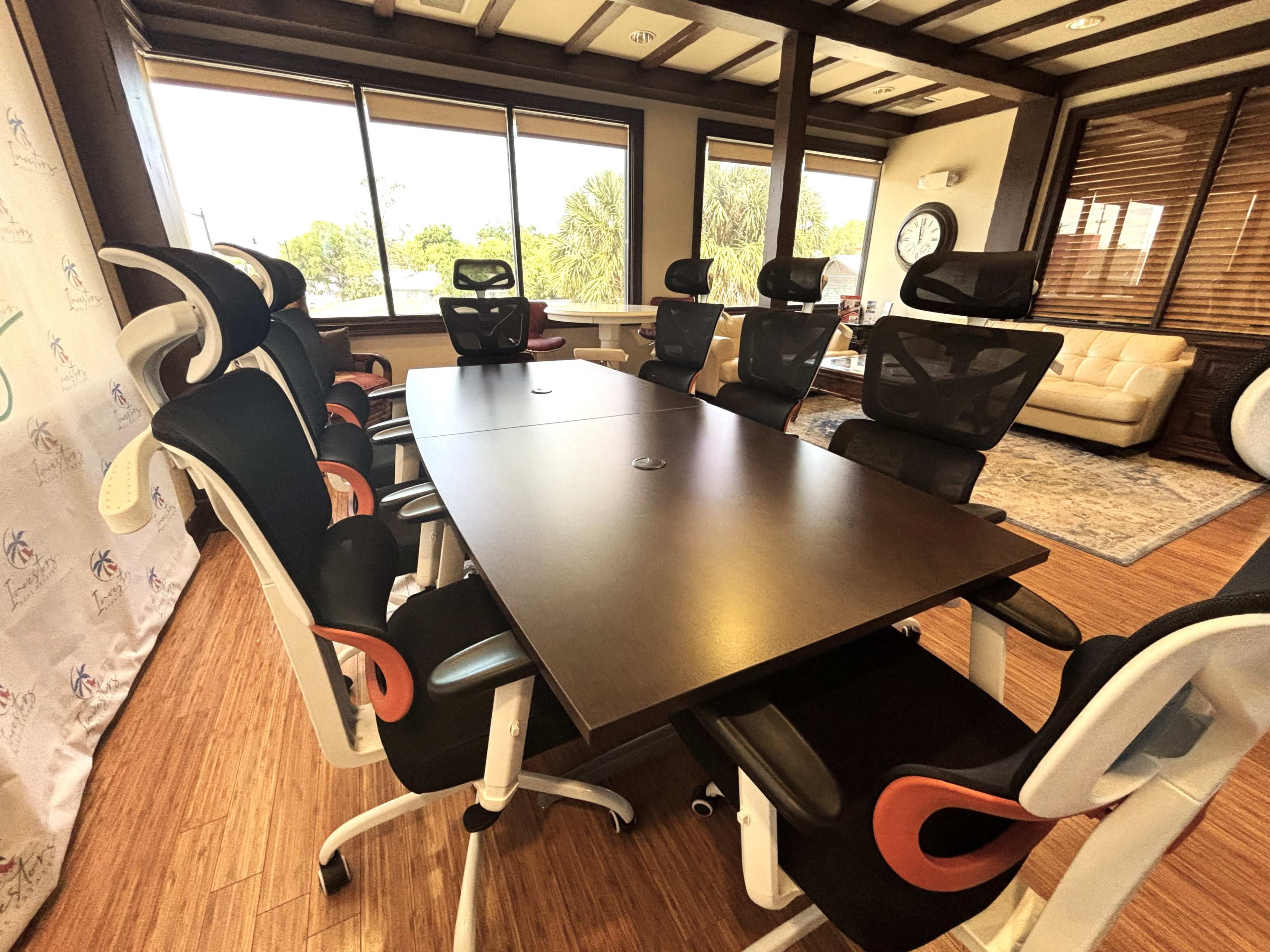 The image shows a modern conference room with a long wooden table surrounded by ergonomic chairs, large windows, and a comfortable seating area in the background.