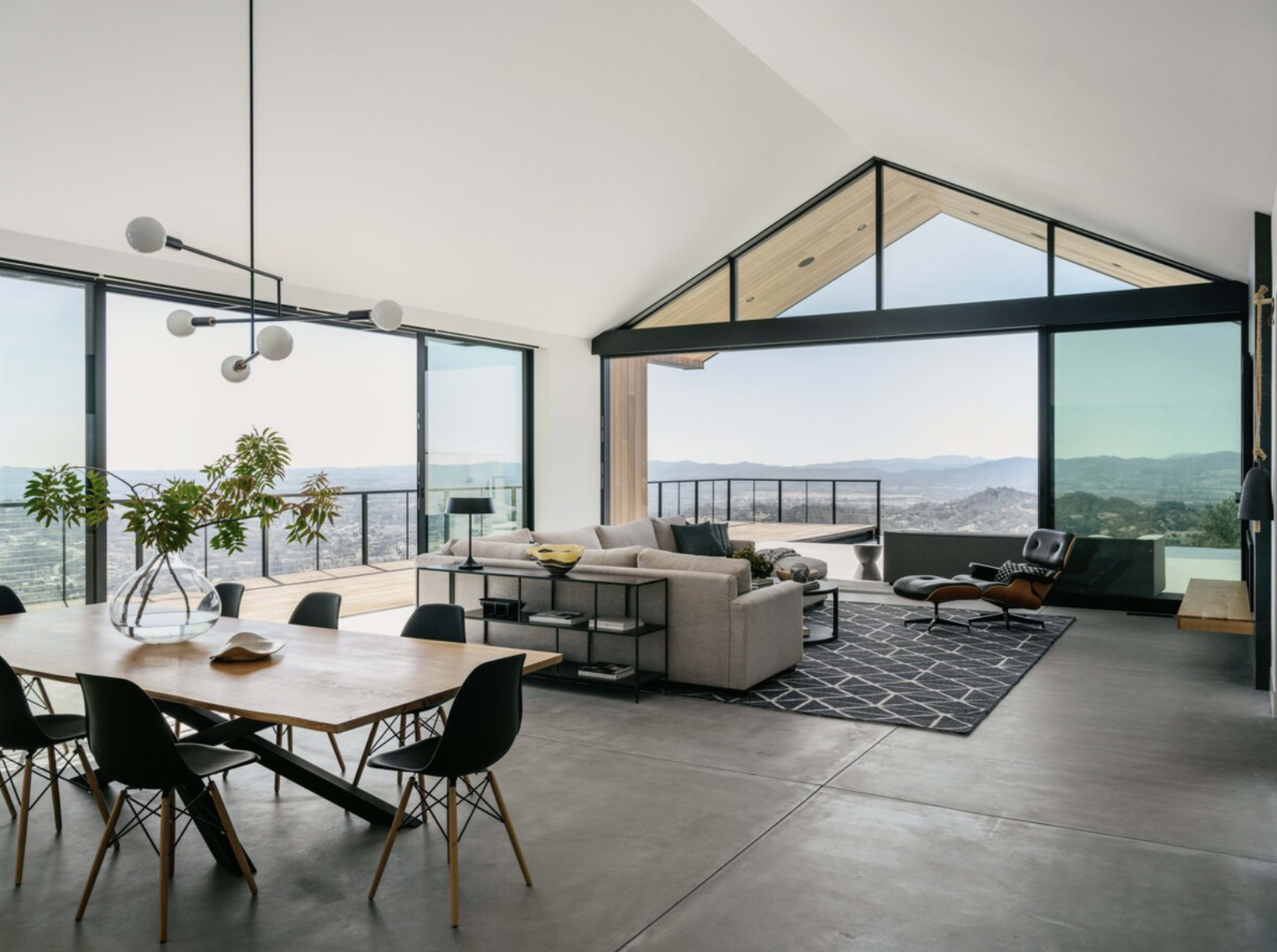 A modern living space with a dining area, a sofa, and large windows offering a view of a distant landscape.