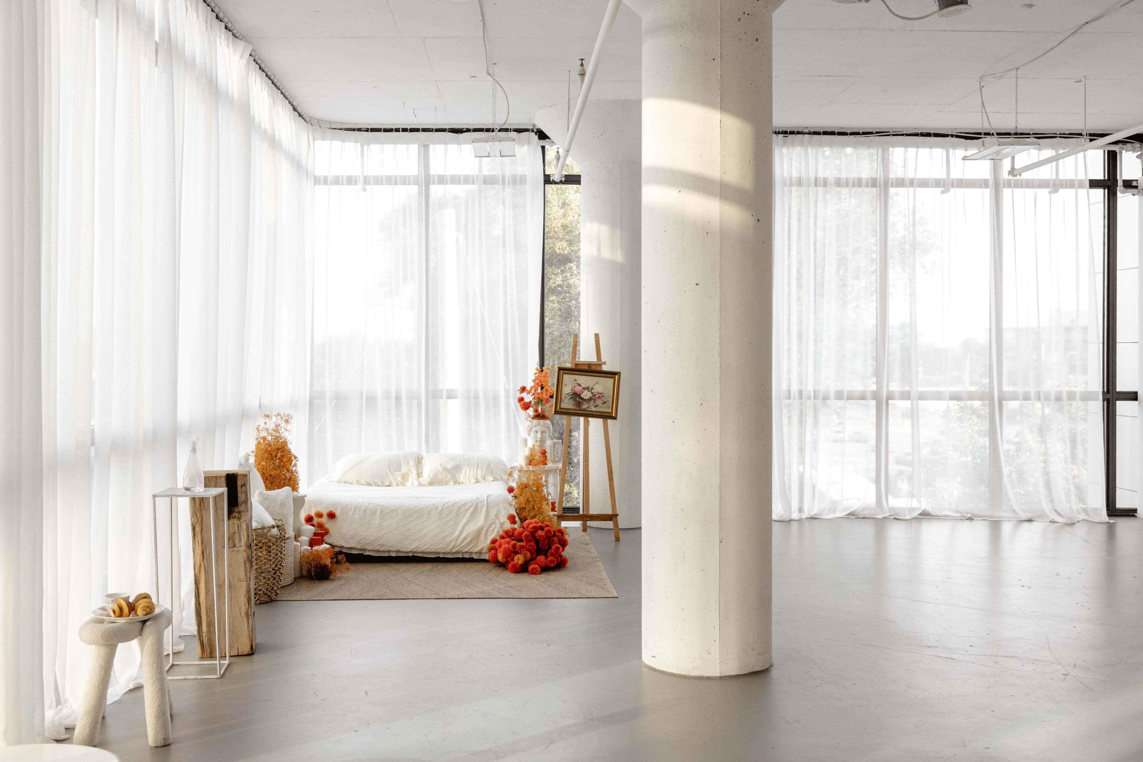The image depicts a modern, minimalist bedroom with large windows covered by sheer curtains, featuring a bed adorned with soft linens and decorative elements such as orange flowers and a small easel displaying artwork.