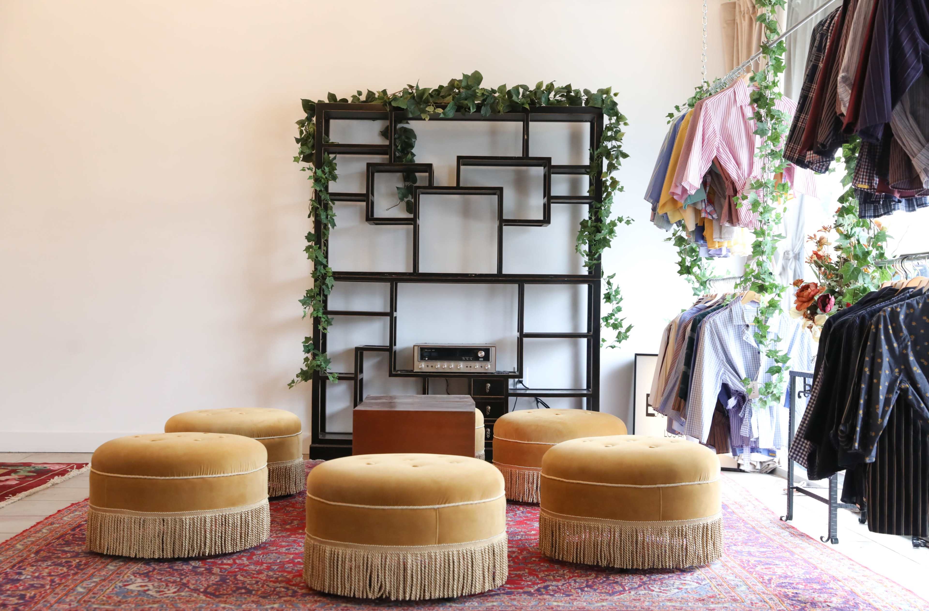 A boutique interior featuring a display shelving unit with greenery, five round ottomans, and clothing hanging on a rack.