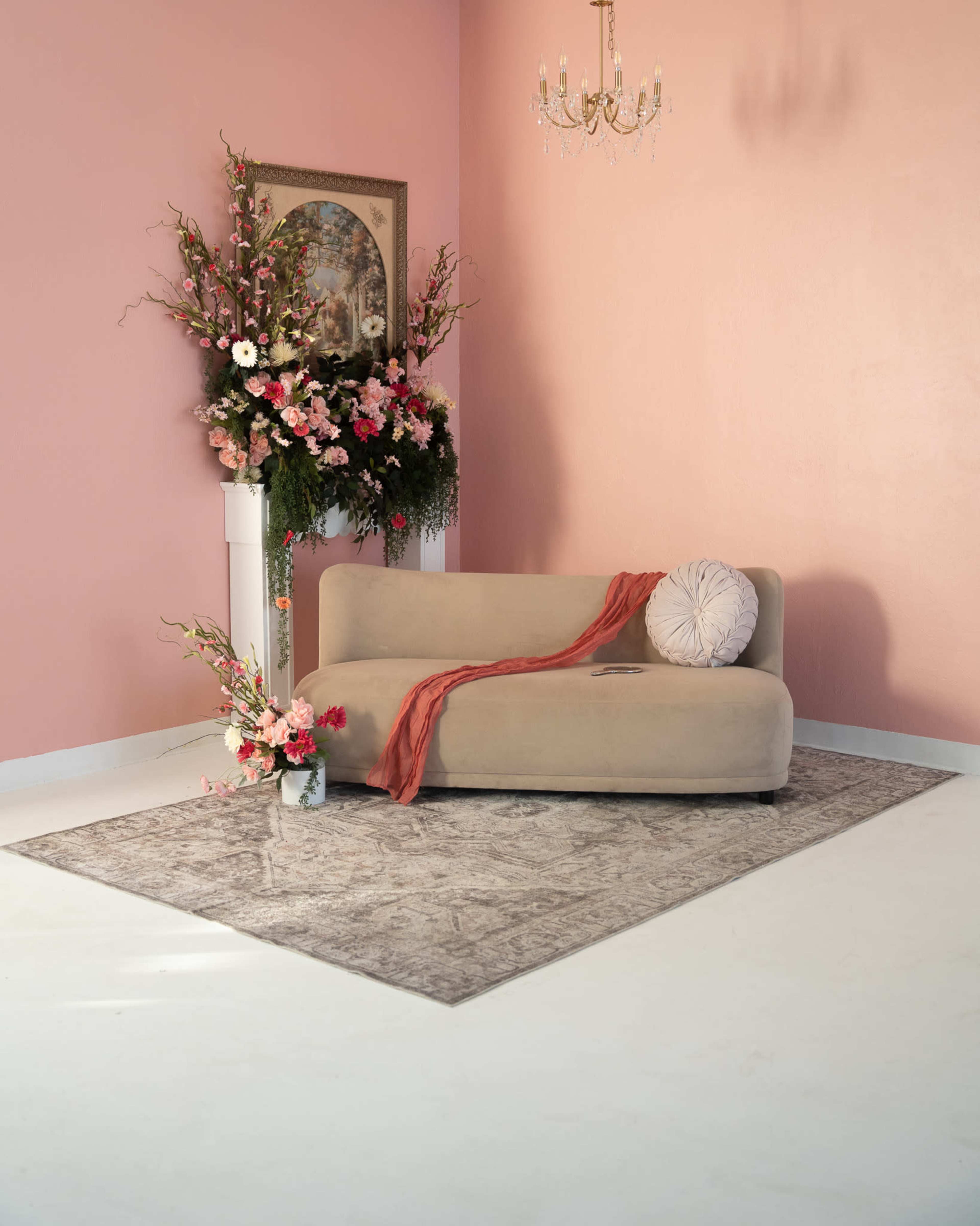 The scene features a light-colored sofa adorned with a decorative cushion, positioned in a softly lit room with a pink wall, a floral arrangement, and a chandelier above.