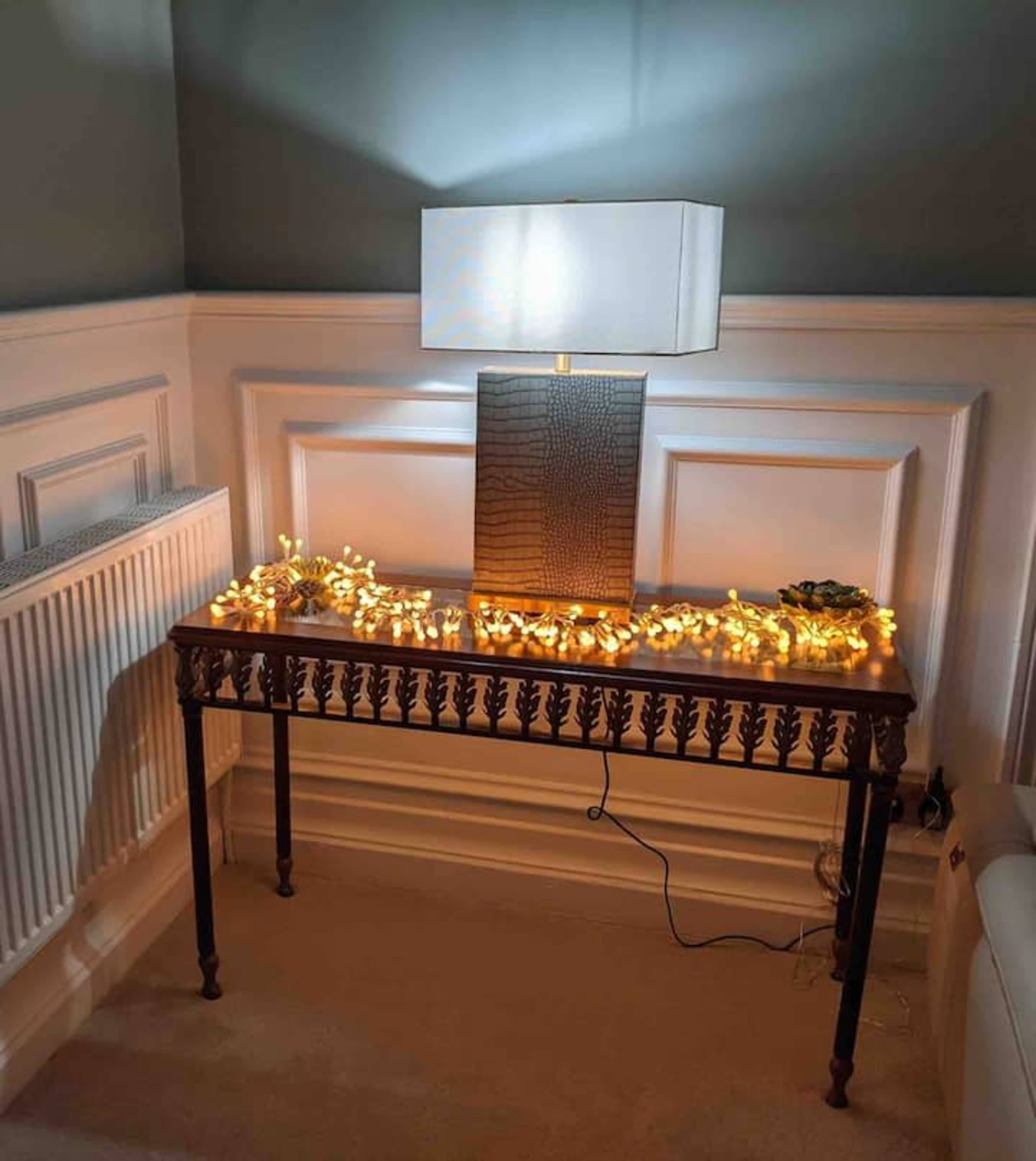 A wooden console table with decorative fairy lights and a rectangular lamp is positioned against a wall with wainscoting.
