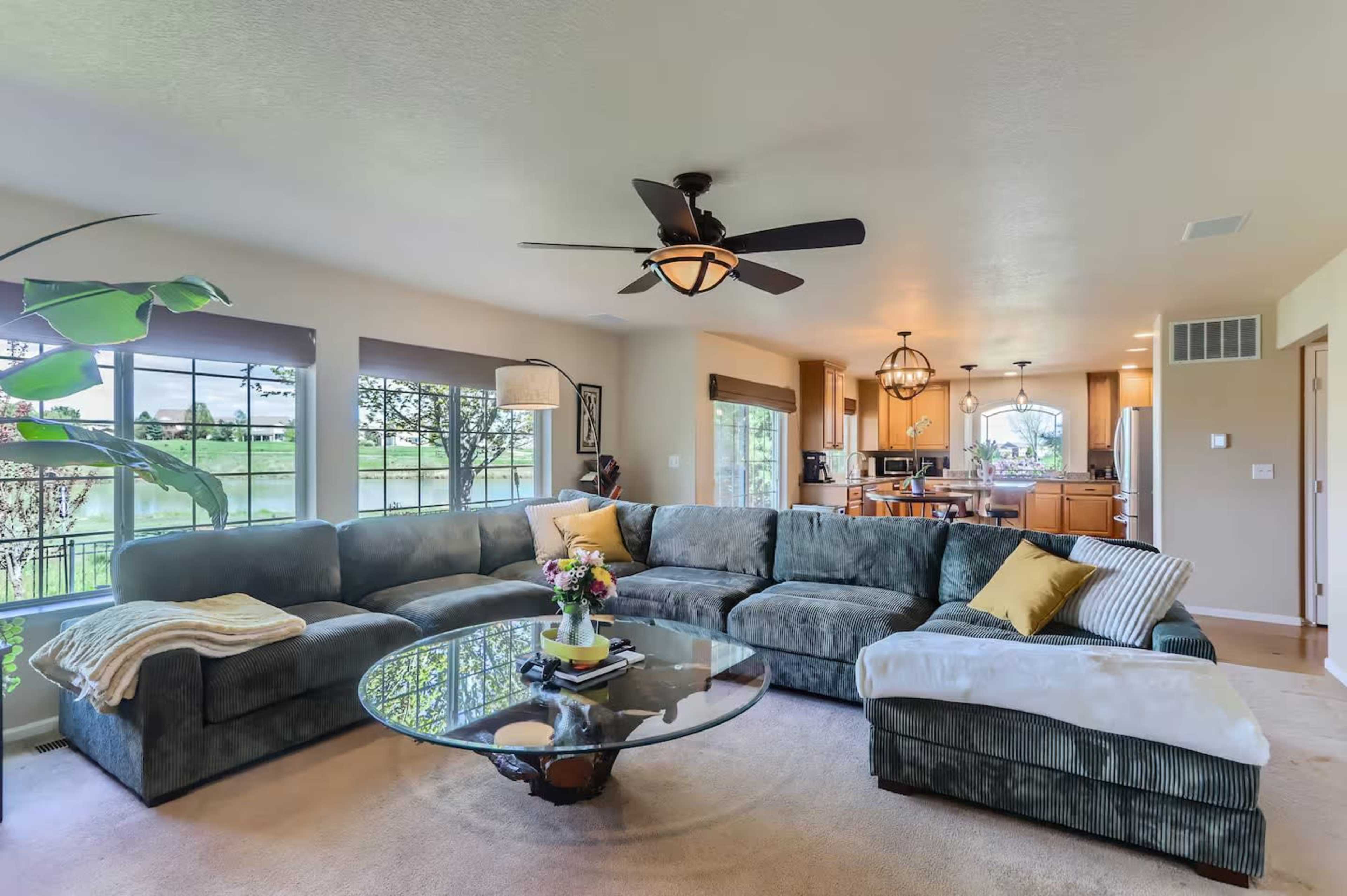 The living room features a large sectional sofa, a glass coffee table, and large windows that provide a view of the outdoor landscape.