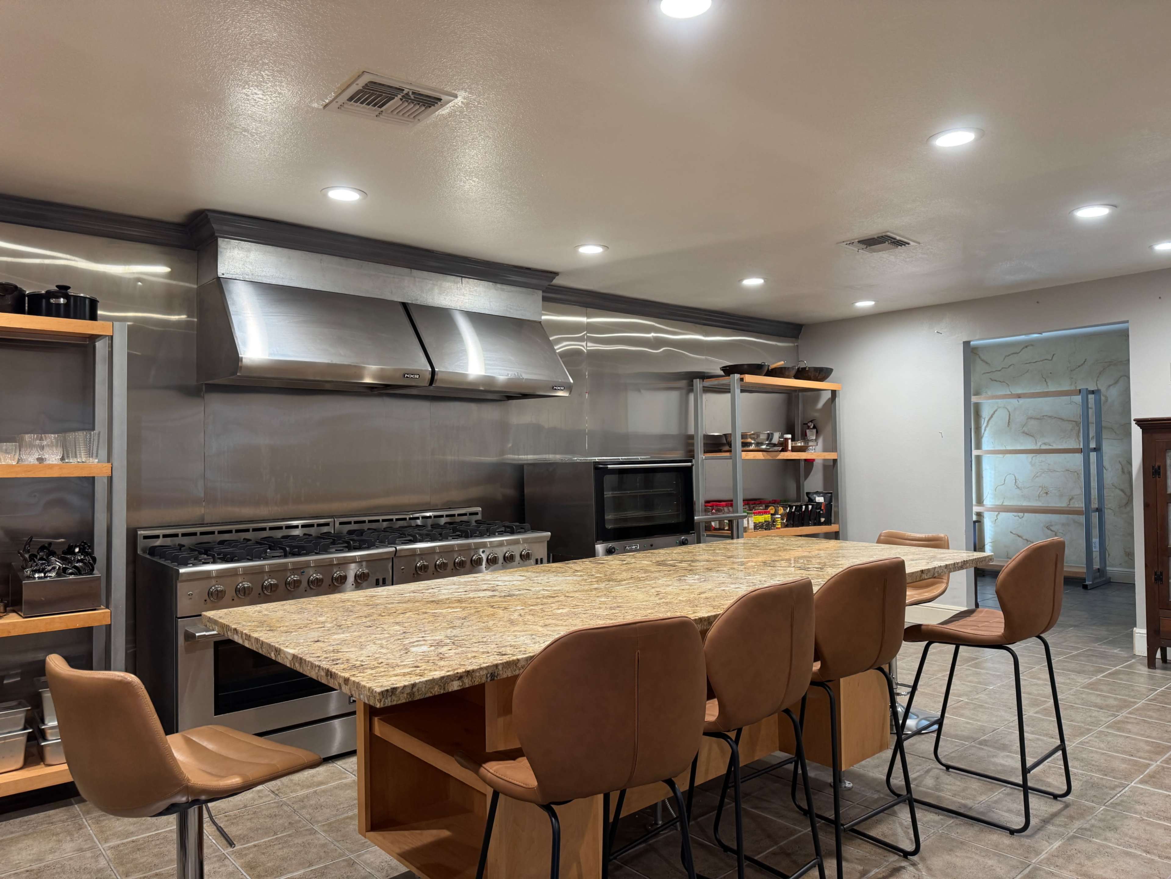 The image shows a modern kitchen with stainless steel appliances, a large granite island with seven brown chairs, and wooden storage shelves.