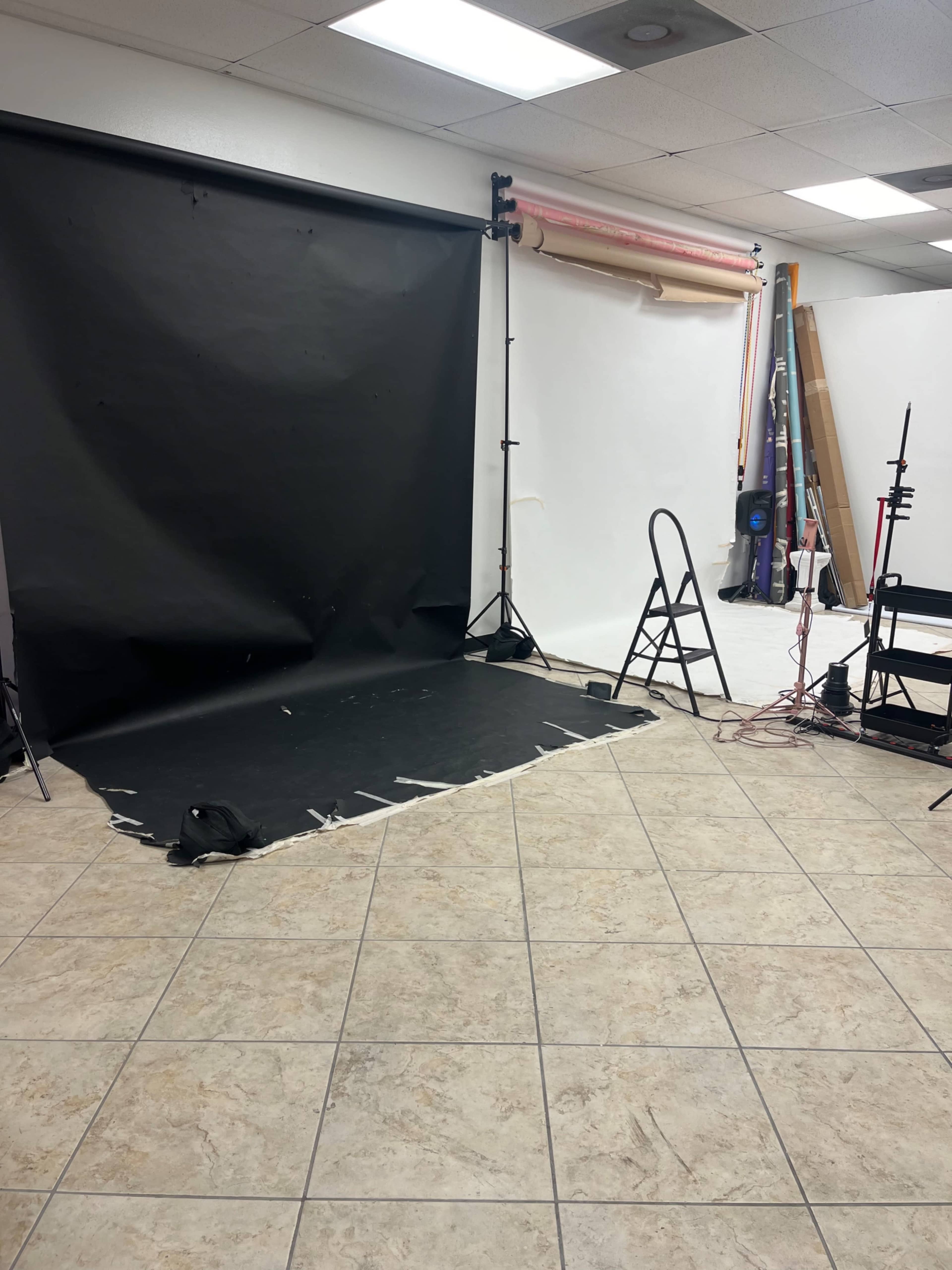 The image shows a photography studio with a black backdrop, a light stand, and a wooden ladder positioned on a tiled floor.