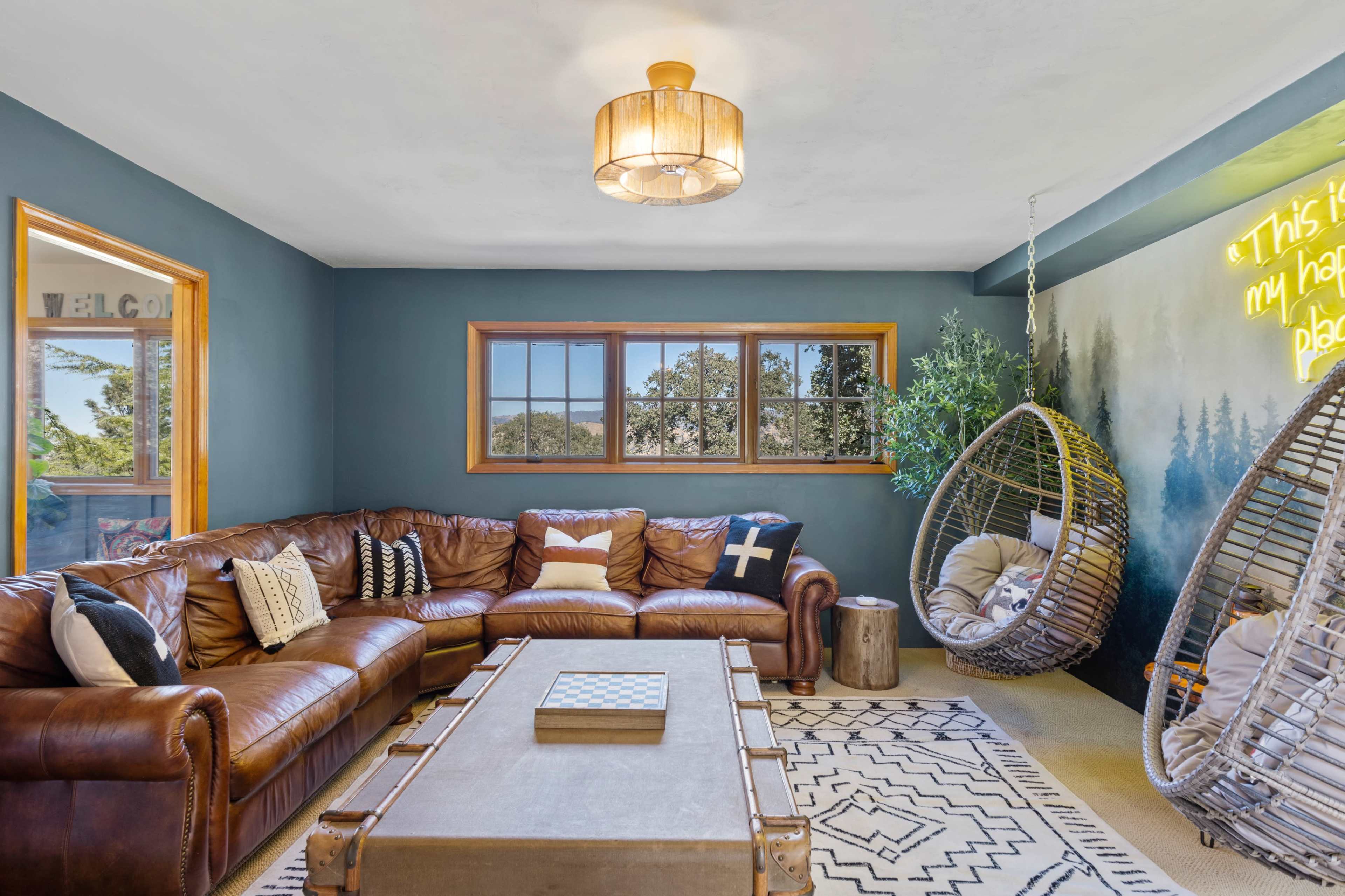 A cozy living room features a large brown leather sofa, two hanging chairs, and a coffee table, with green plants and a window showcasing outdoor scenery.