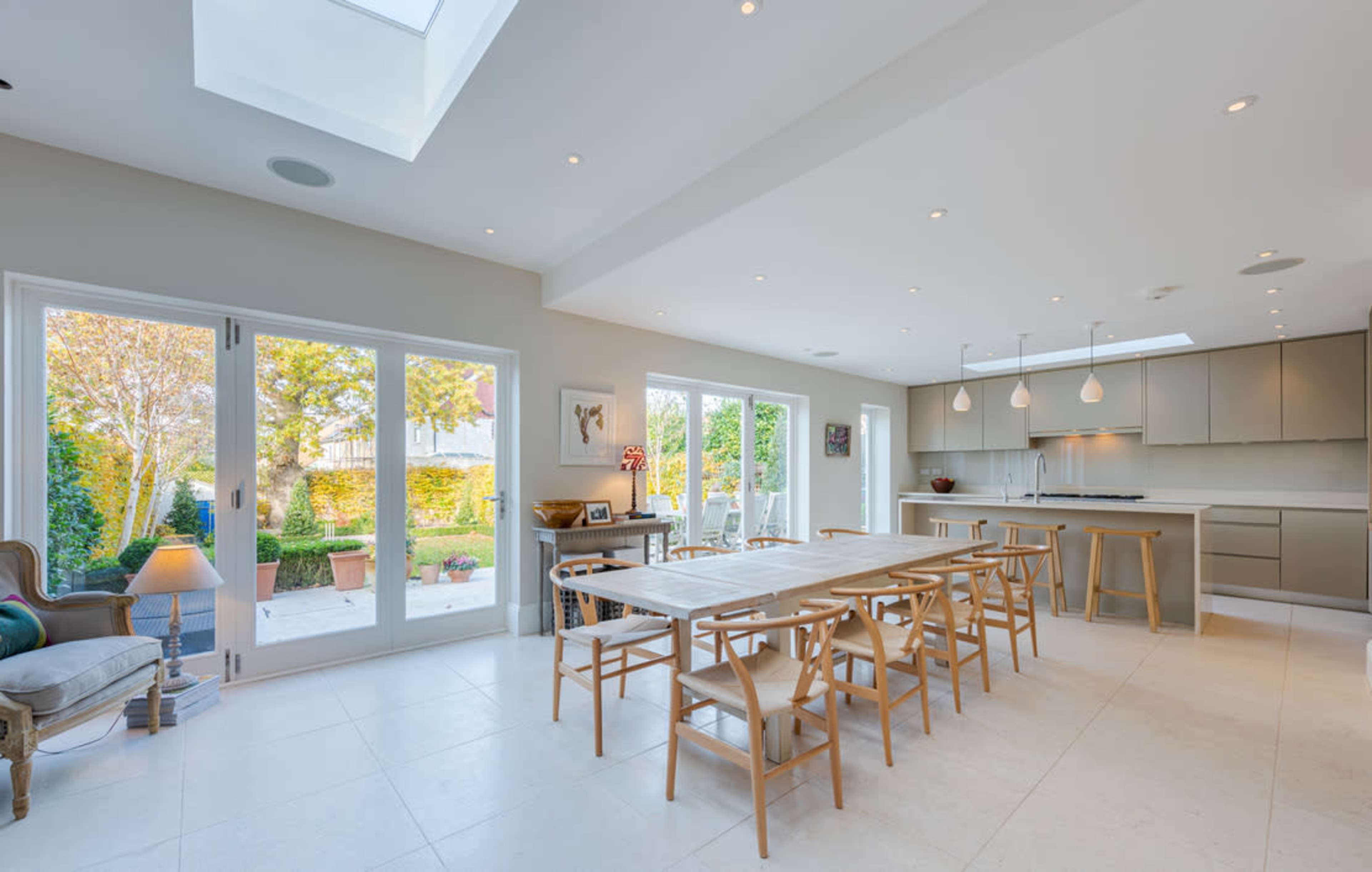 A modern kitchen and dining area with a large table, wooden stools, and glass doors opening to a garden.