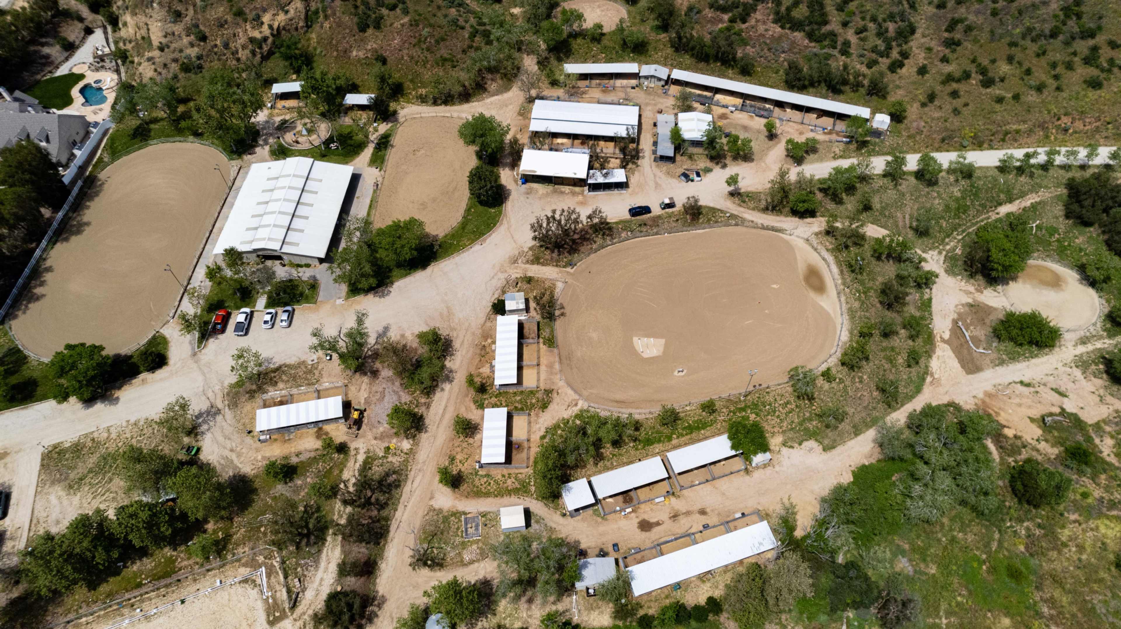 The aerial view shows a horse training facility with several buildings, riding arenas, and a pool area surrounded by trees.