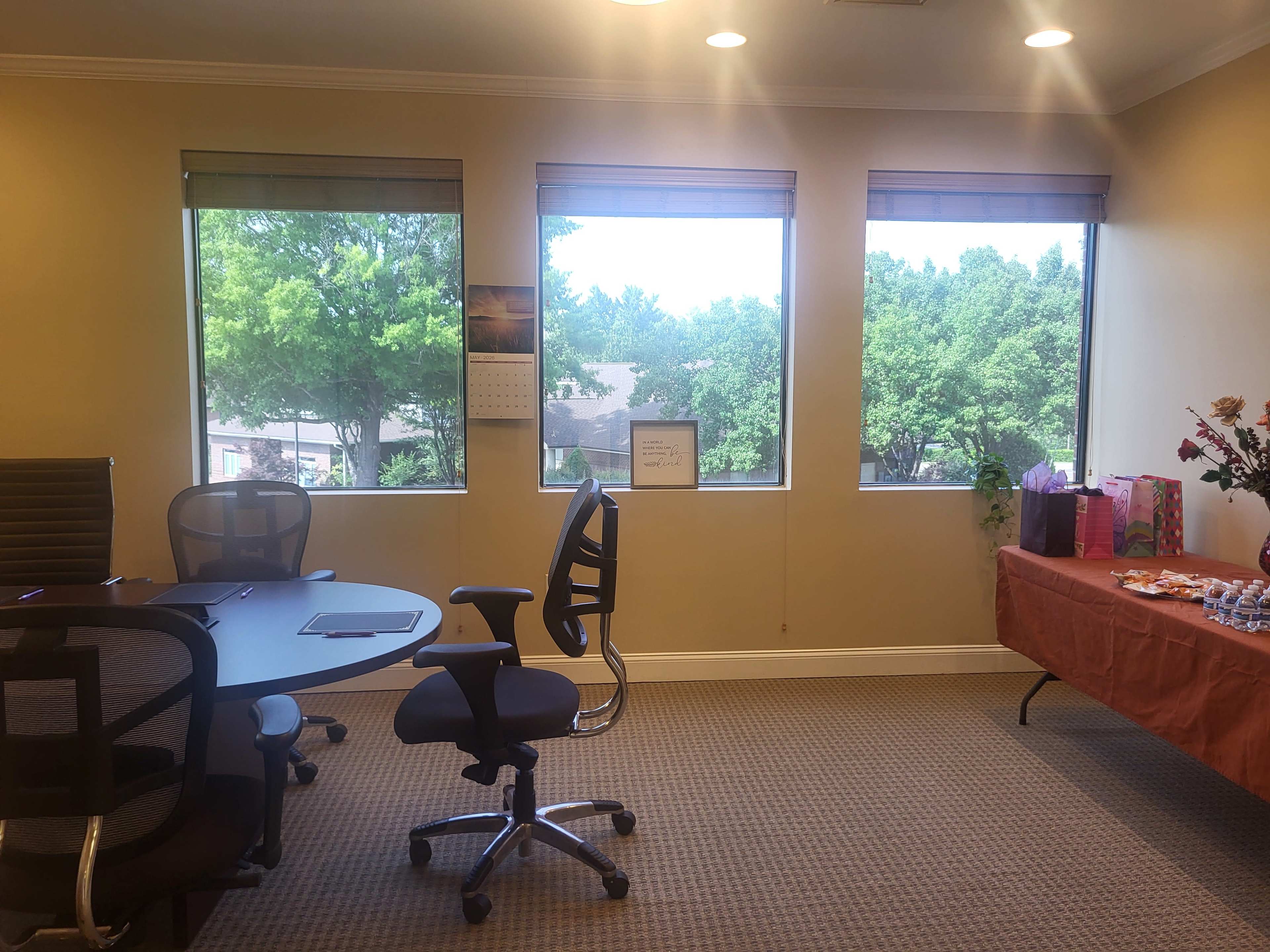 The image shows a bright office space with three large windows, a round table with chairs, and a side table decorated for a small gathering.
