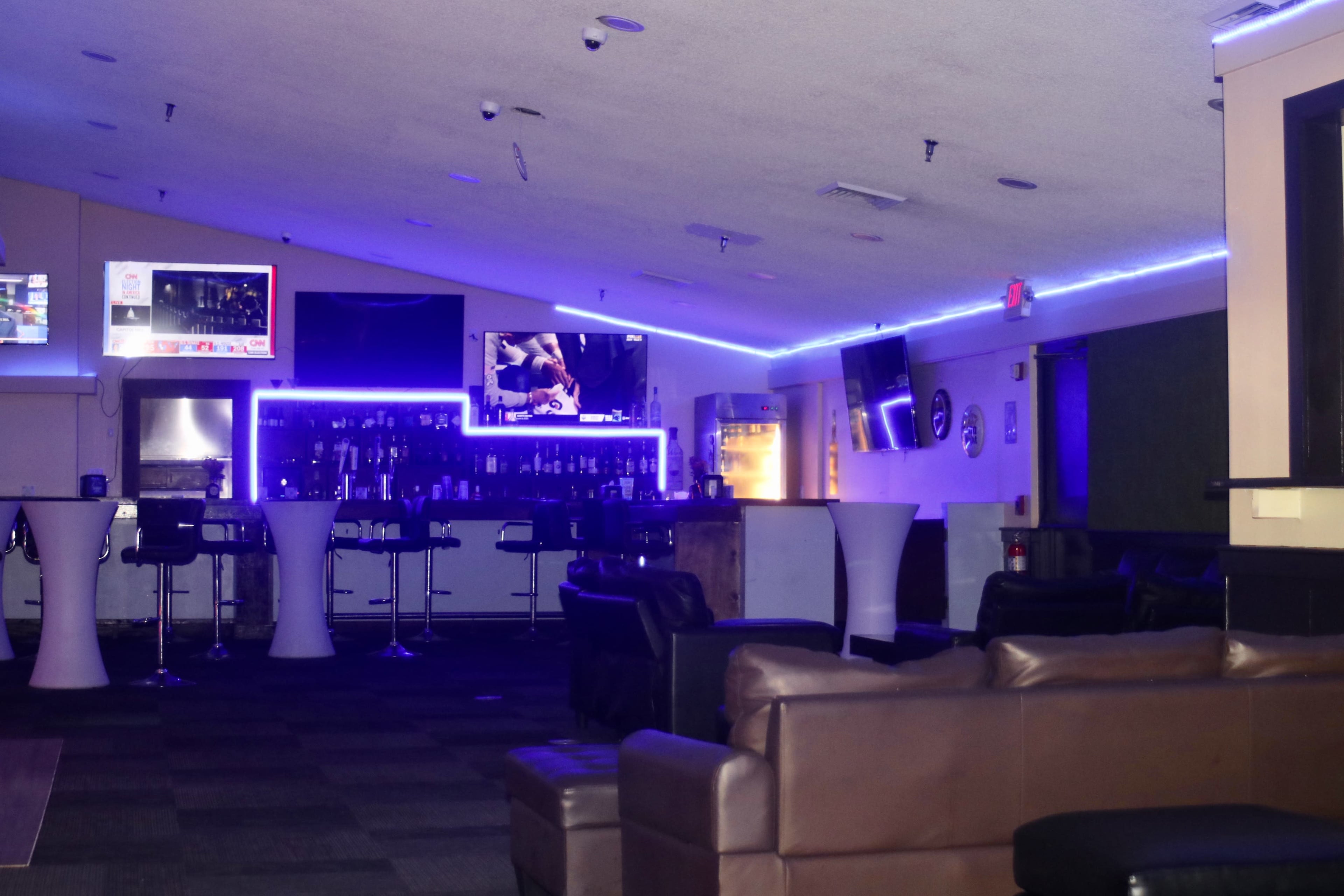 The image shows an empty lounge area with a bar featuring illuminated shelves, several television screens, and modern seating arrangements.