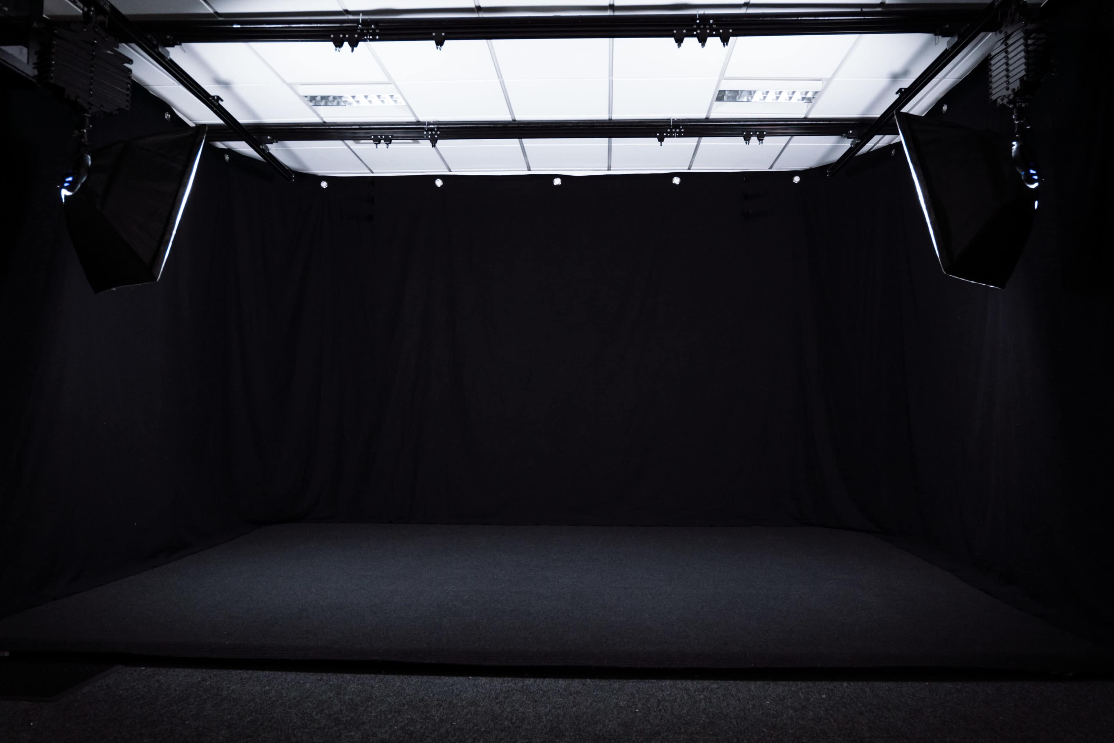 A dimly lit studio space with black curtains and a dark carpeted floor, and softbox lights mounted above.