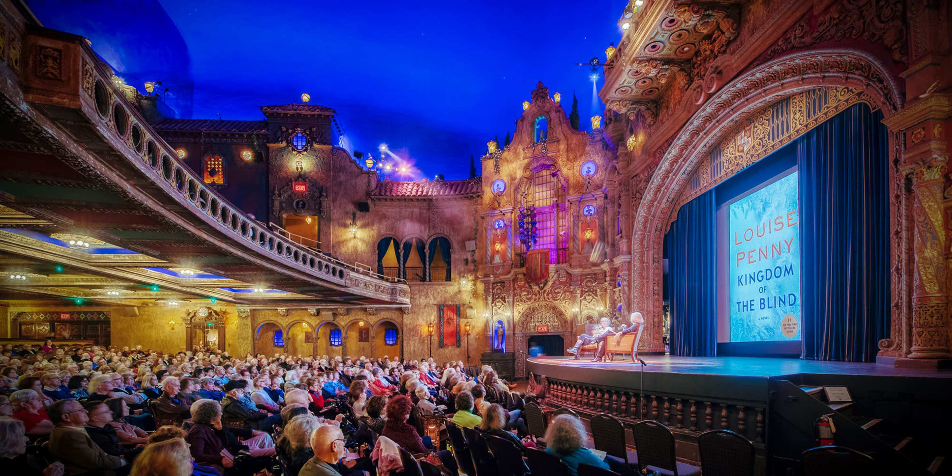A large audience sits in a historic theater with ornate decorations, facing a stage featuring a presentation about Louise Penny's book "Kingdom of the Blind."