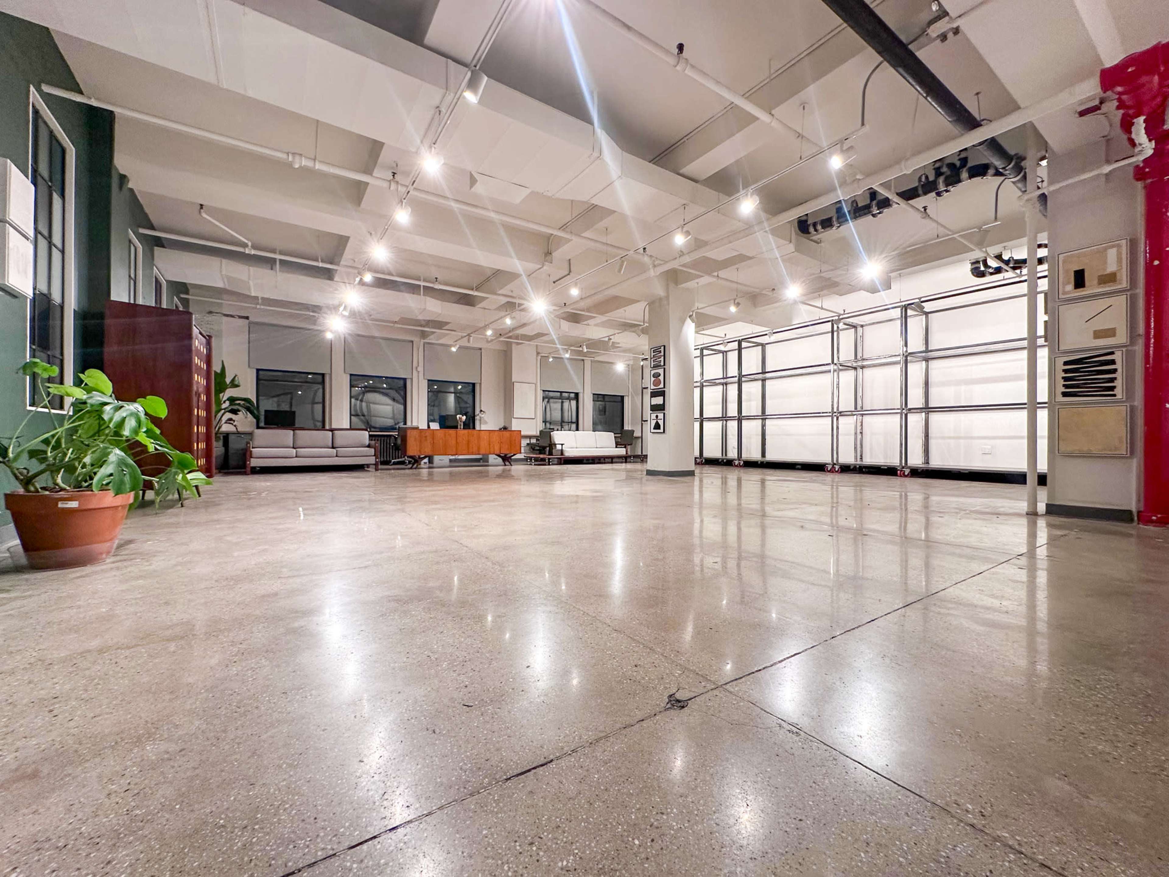 The image shows a spacious, well-lit interior with polished concrete floors, large windows, and furniture arranged in a lounge area.