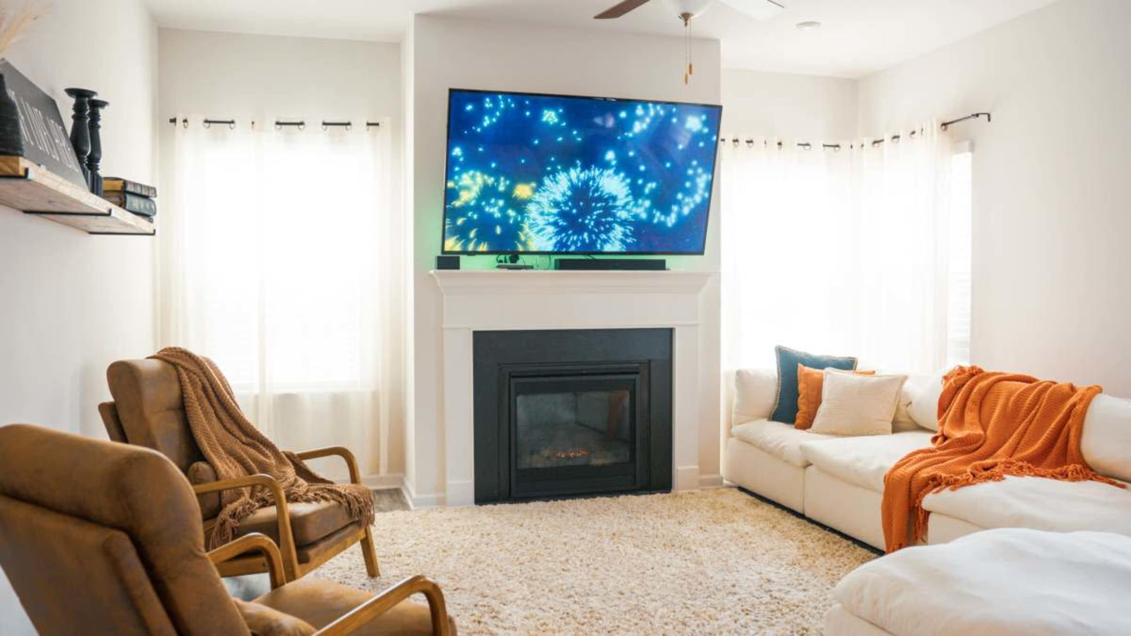The image shows a cozy living room with a large television displaying a colorful fireworks display, two armchairs, a white sofa, and a light-colored area rug.