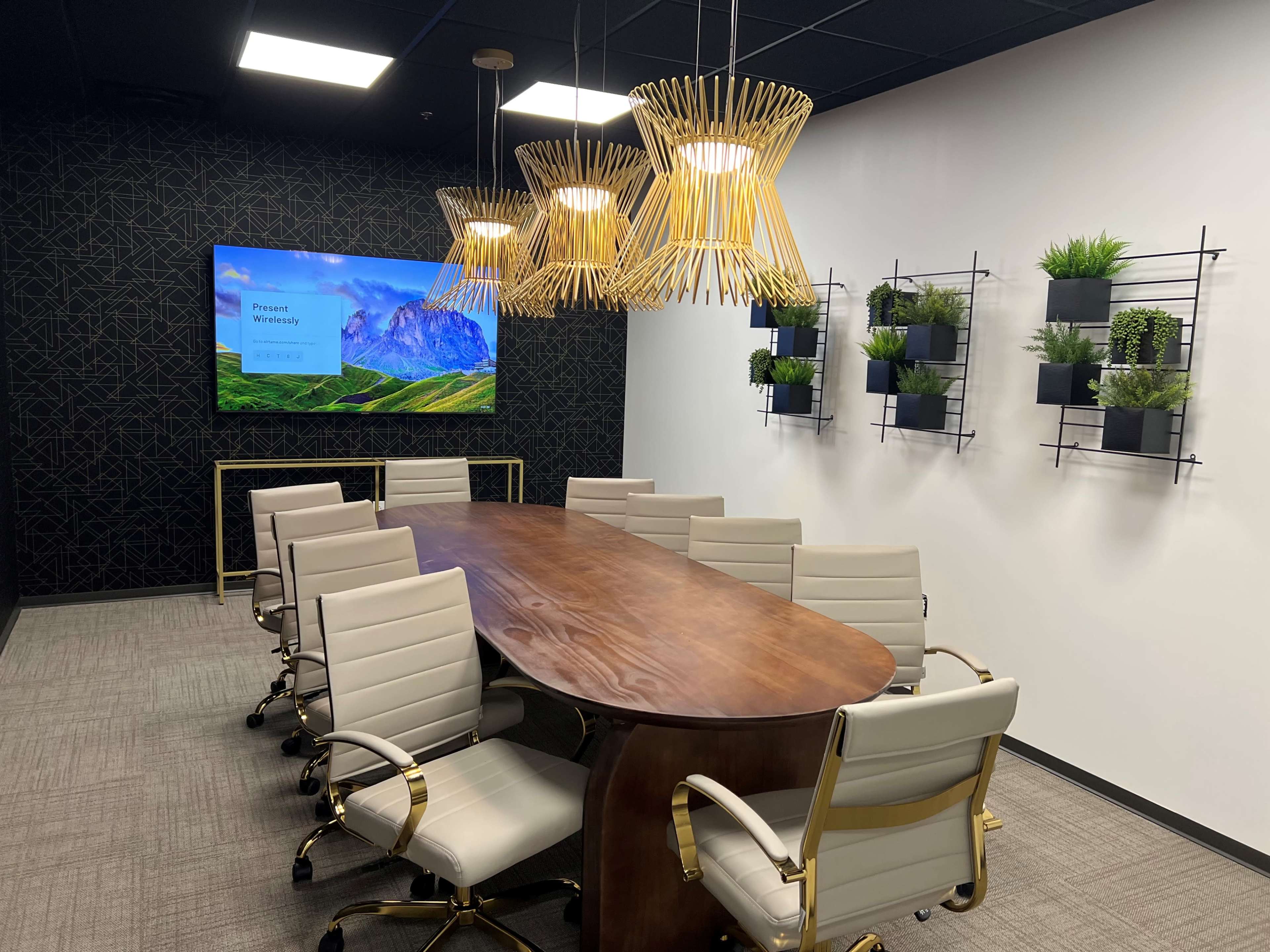 A modern conference room featuring a long wooden table, white chairs, a wall-mounted screen displaying a scenic landscape, and green plants arranged on wall shelves.