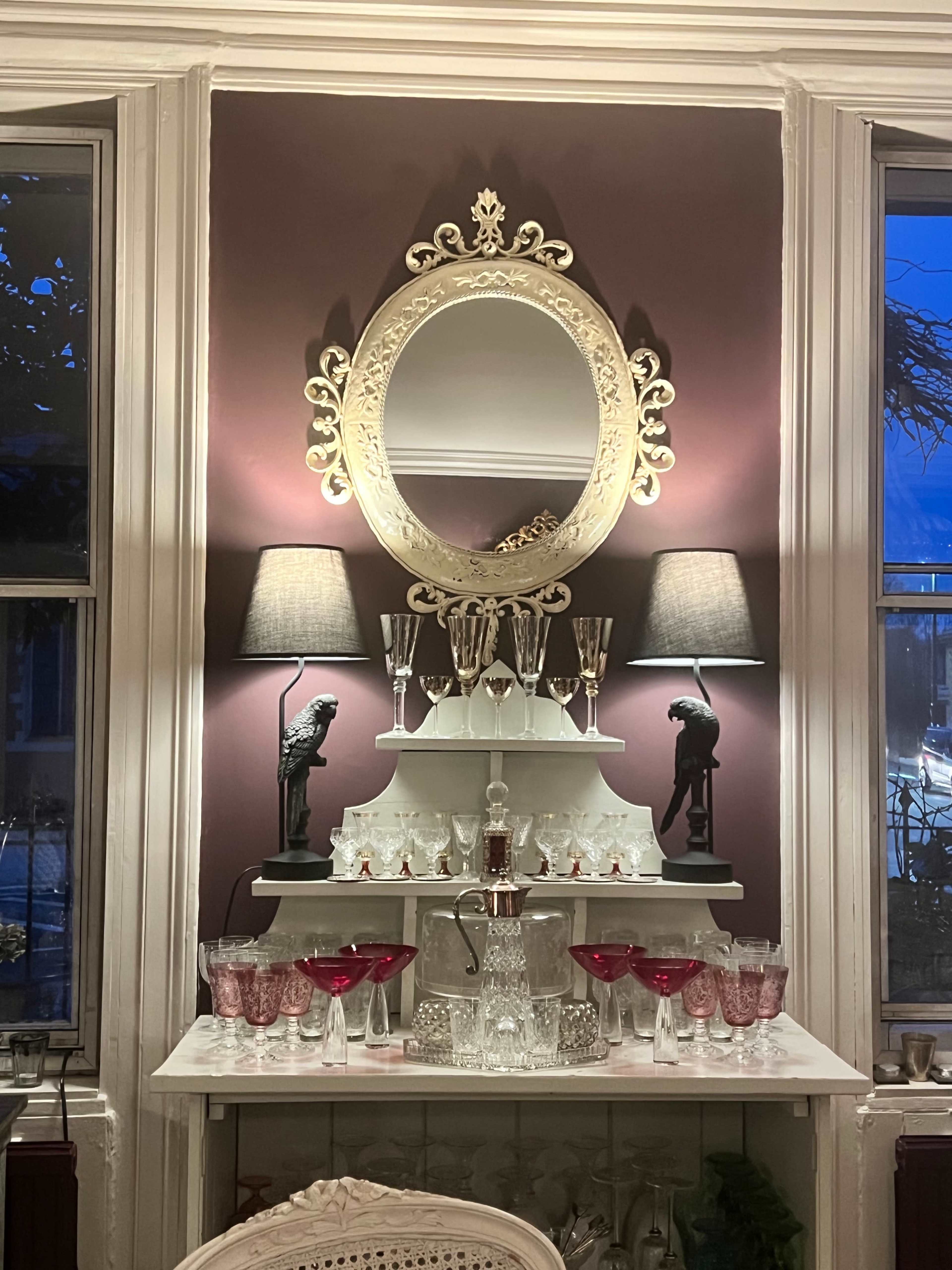A decorative shelf with a large ornate mirror above it, flanked by two lamps, and showcasing an arrangement of glassware, including red and clear crystal glasses, on a white surface.