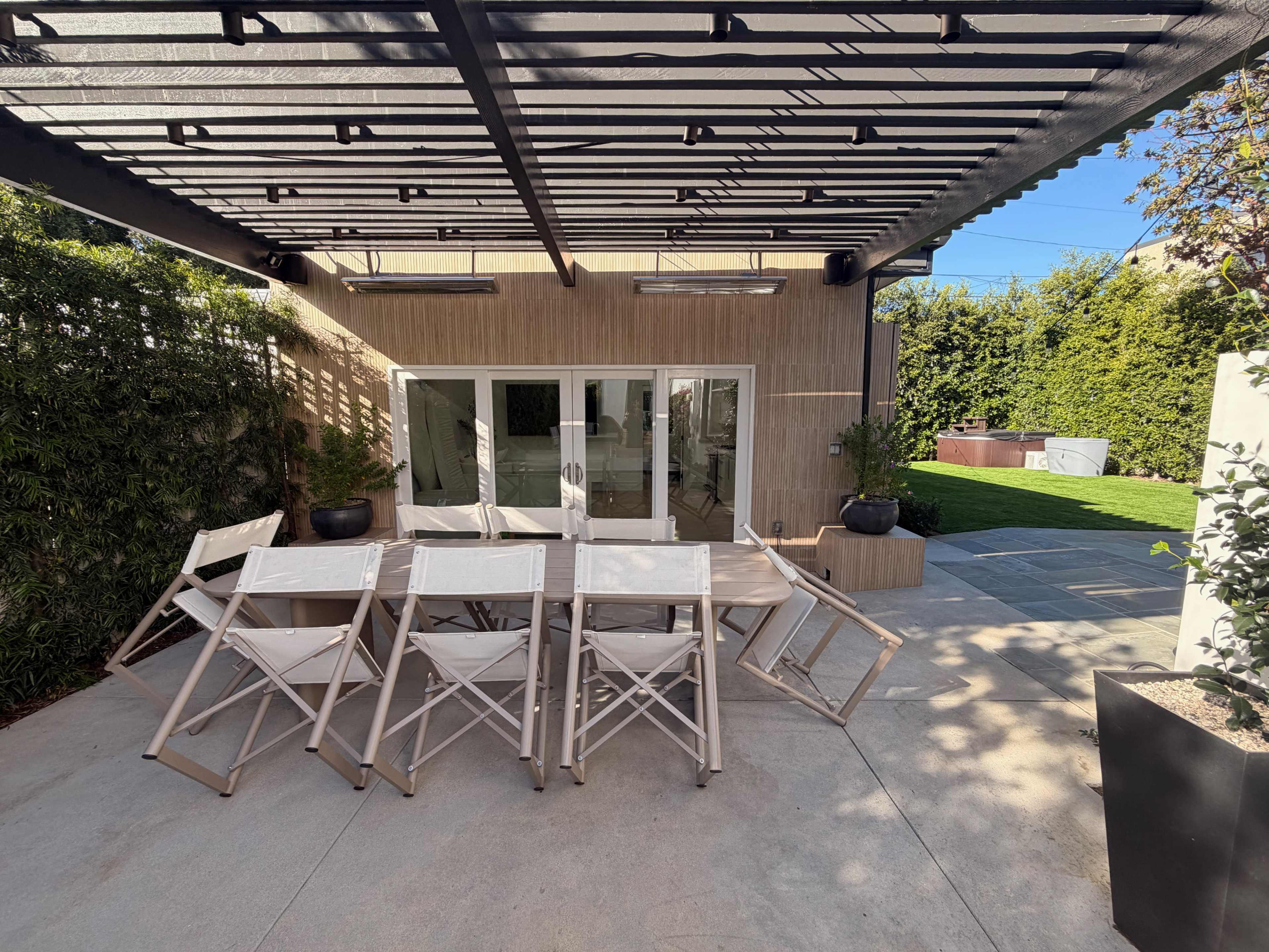 A patio area features a large wooden table surrounded by white folding chairs, with a modern structure overhead, leading to a view of a garden and a glass door.