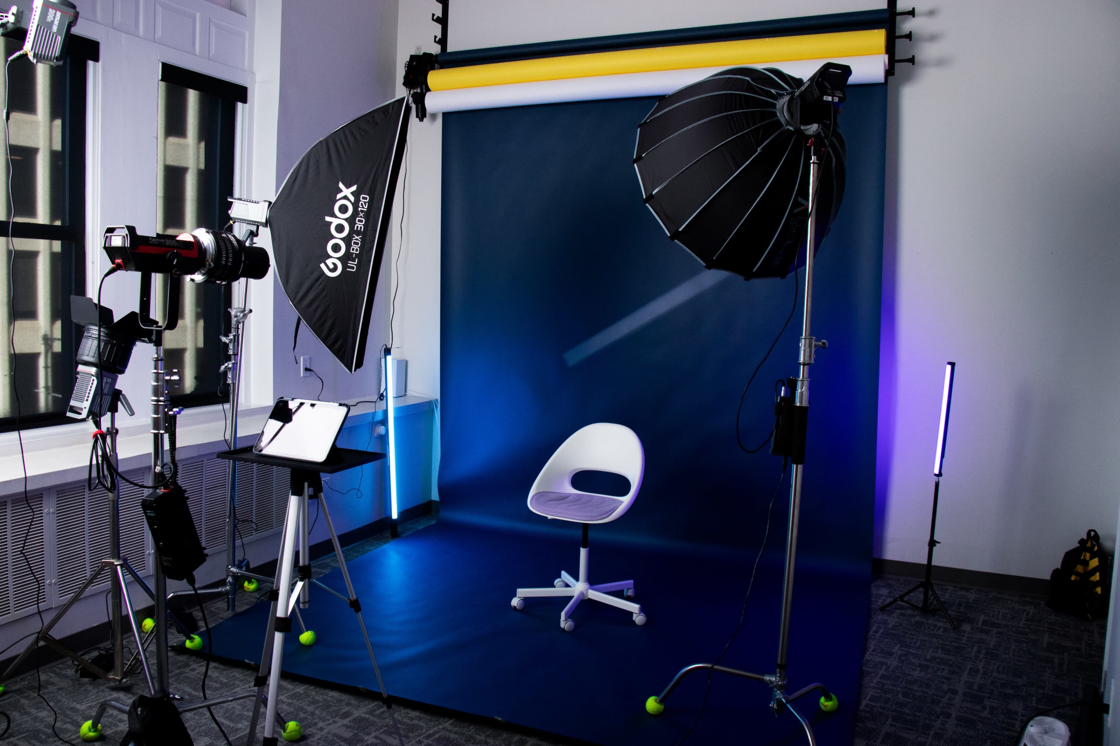 The image shows a photography studio setup featuring a blue backdrop, a chair, and multiple lighting equipment.