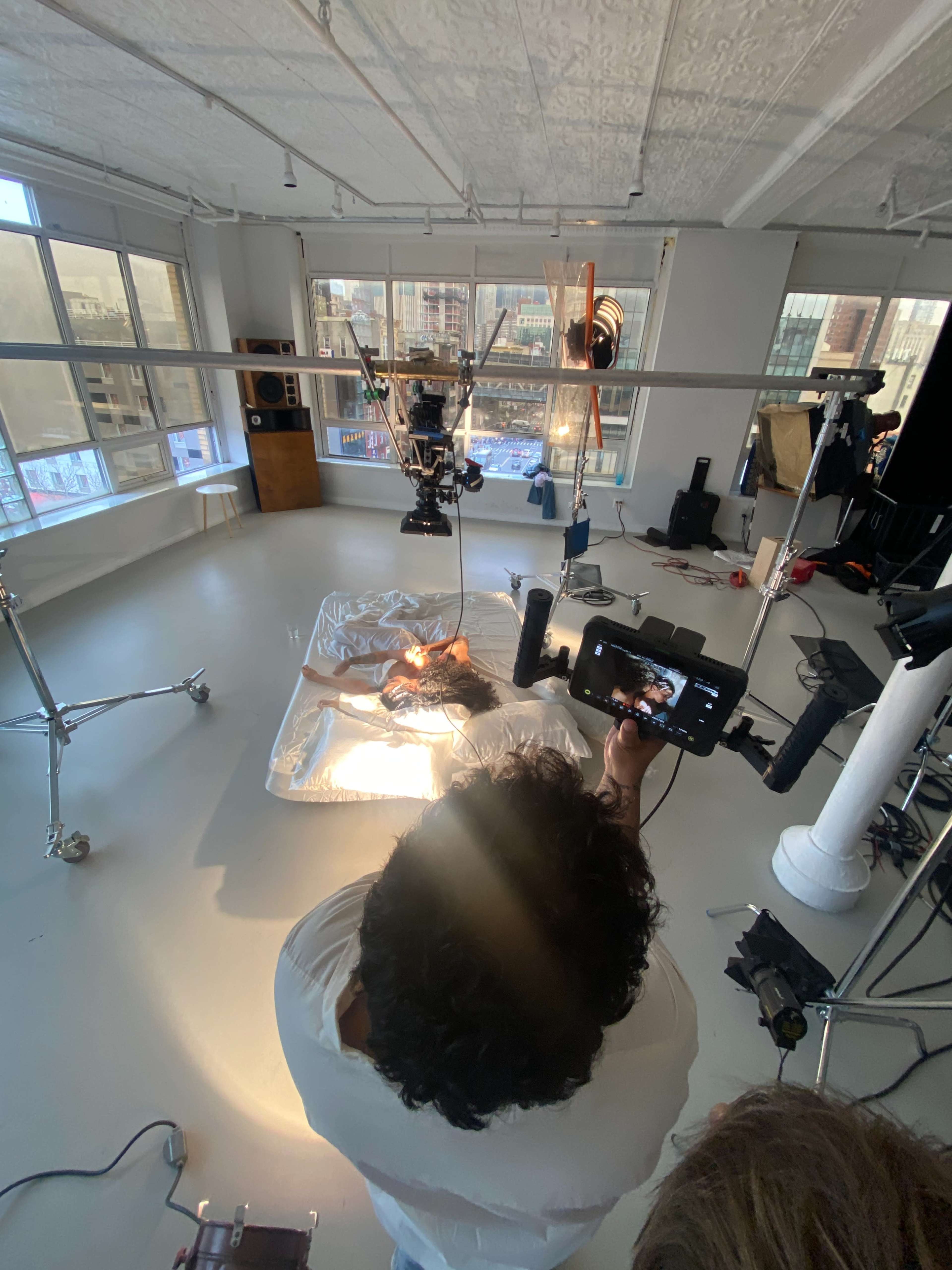A person operates a camera from above a setup with a reflective surface and a subject in a studio filled with various equipment and large windows.