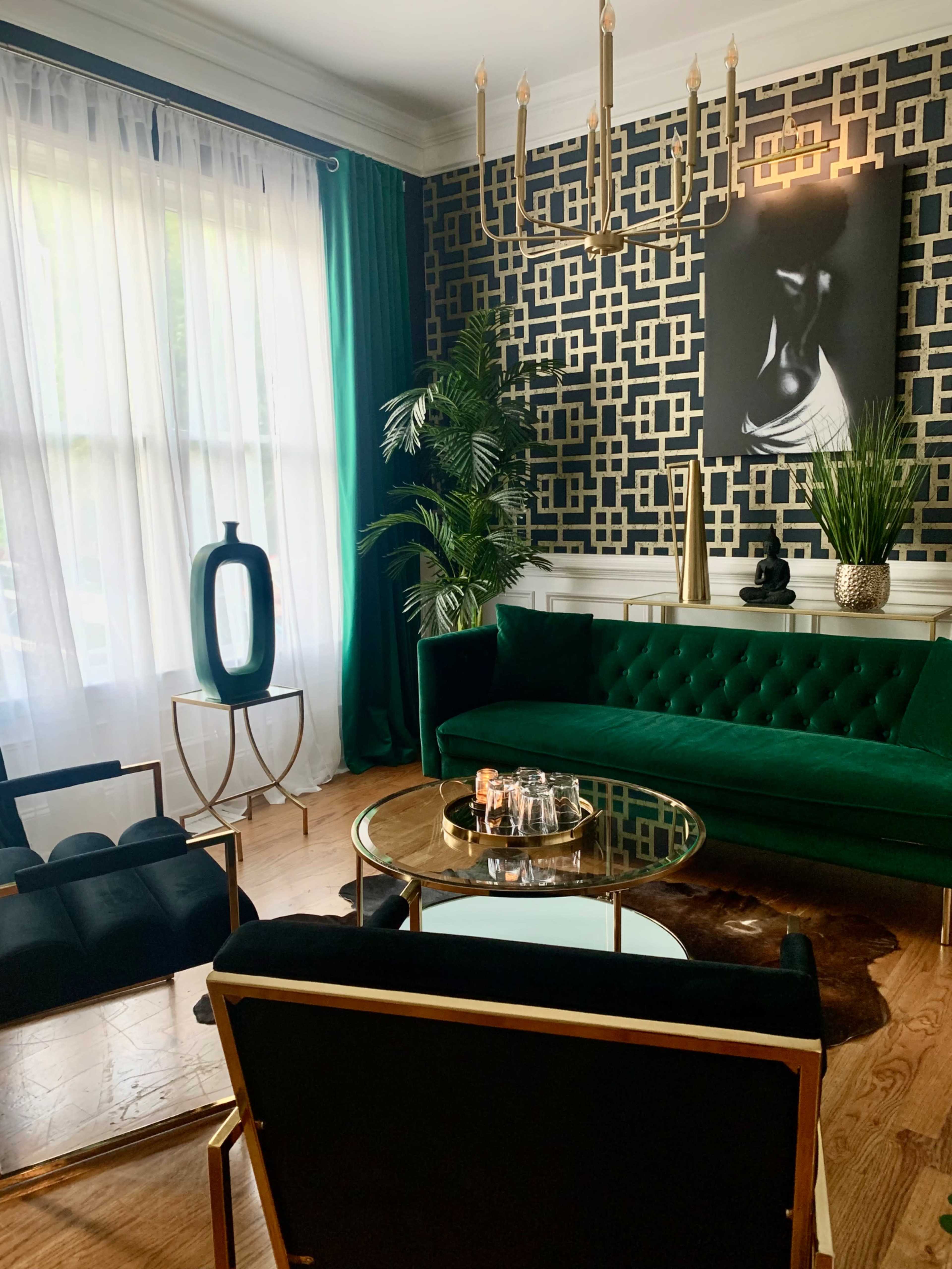 A stylish living room featuring a green velvet sofa, a gold coffee table, and decorative plants against a patterned black and gold wall.