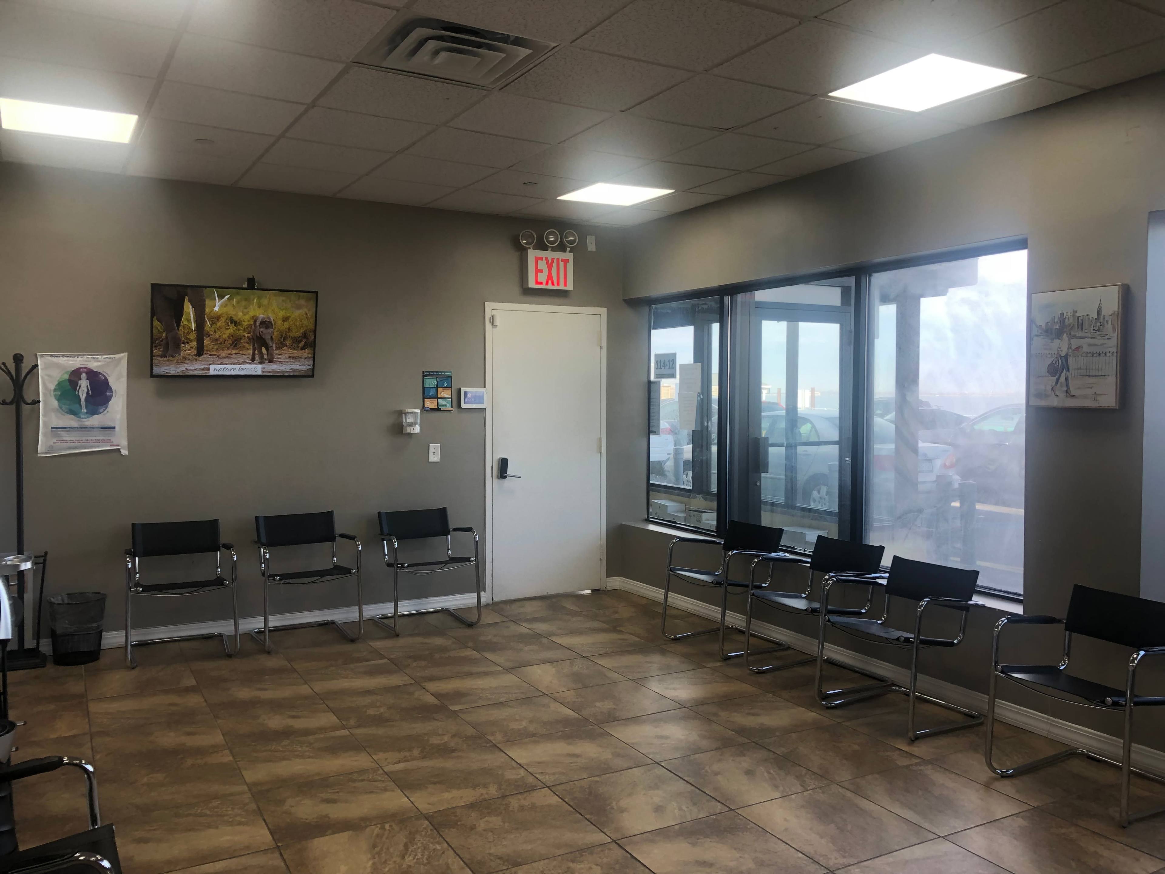 The image shows a waiting room with several metal chairs, a television mounted on the wall, and a door leading outside.