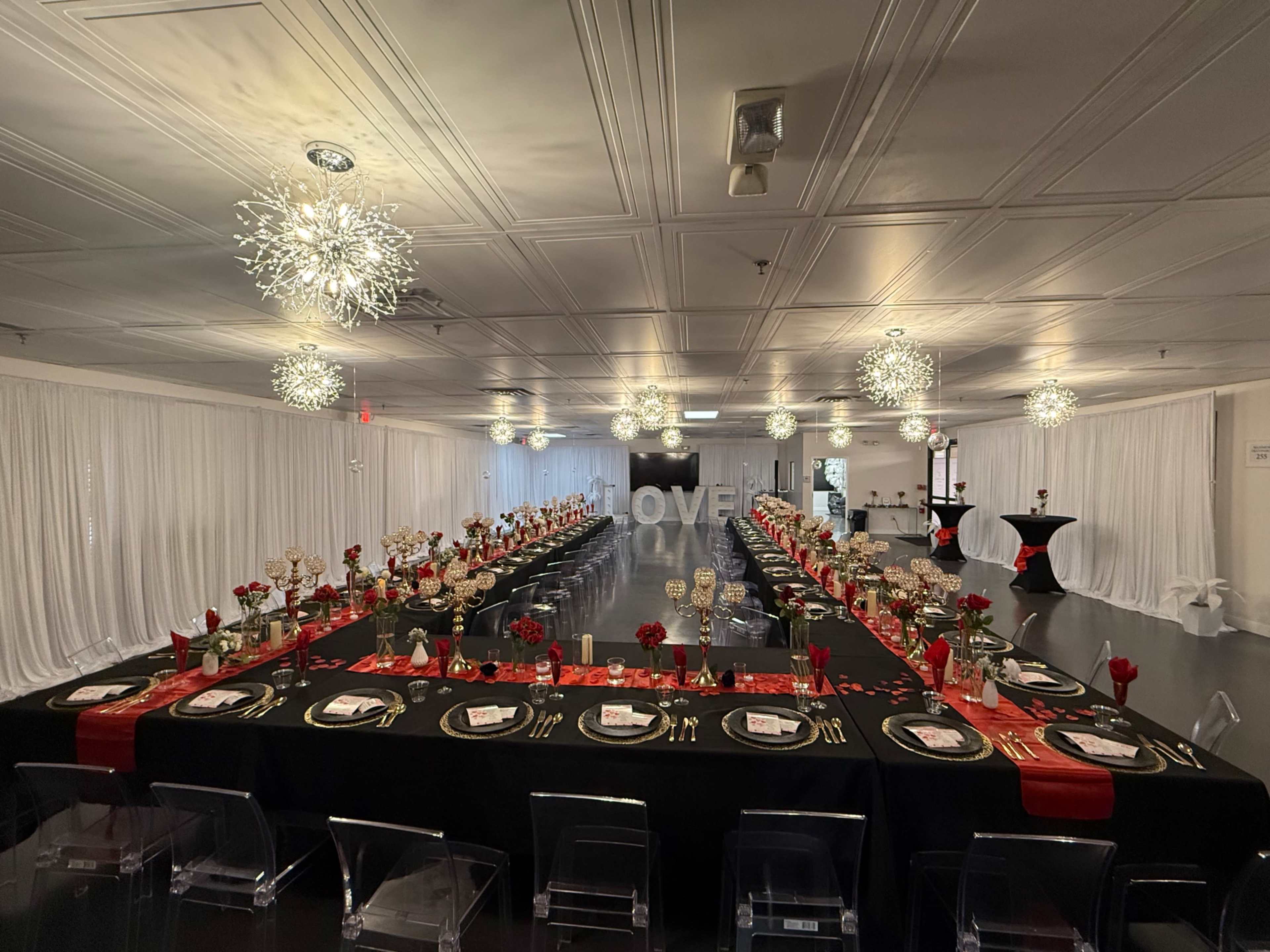 A large banquet hall is arranged with a long table set for a formal dinner, featuring elegant decorations, red roses, and sparkling lighting fixtures.