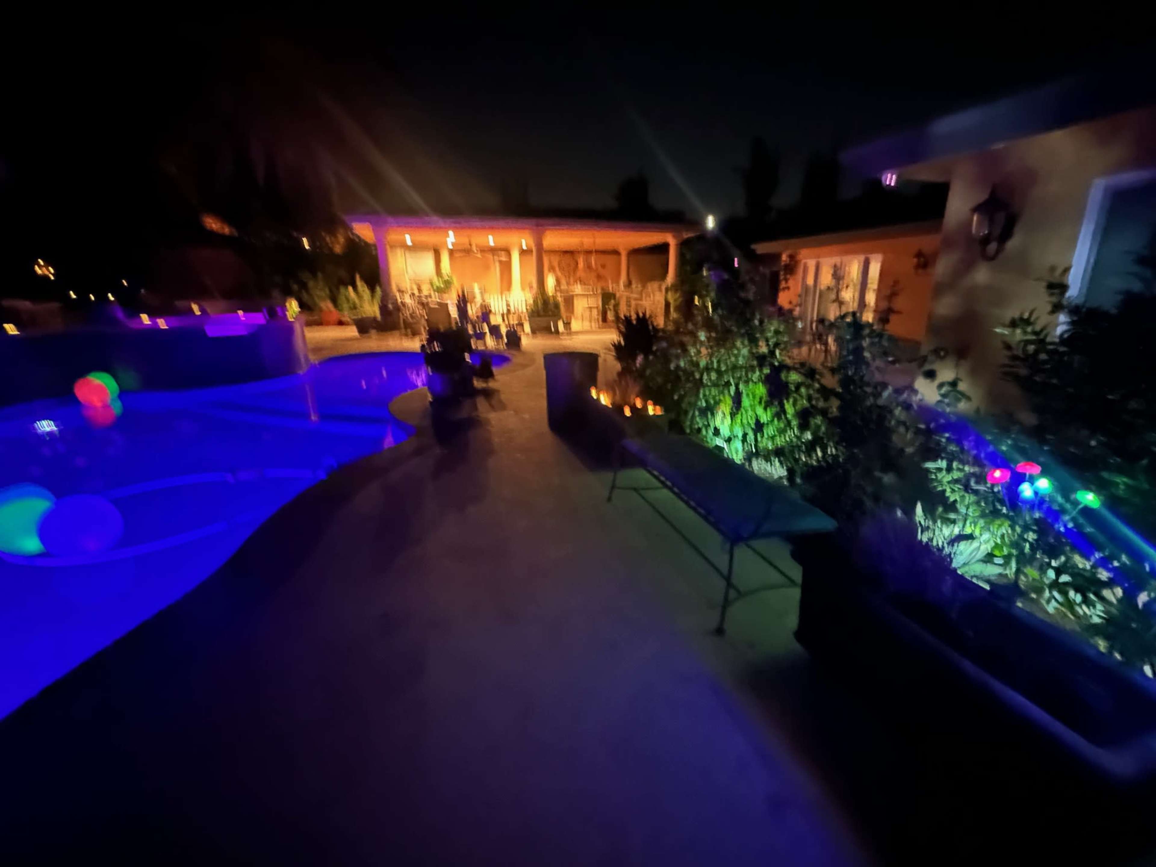 A well-lit backyard features a swimming pool with colorful lights, surrounded by plants and a patio area.