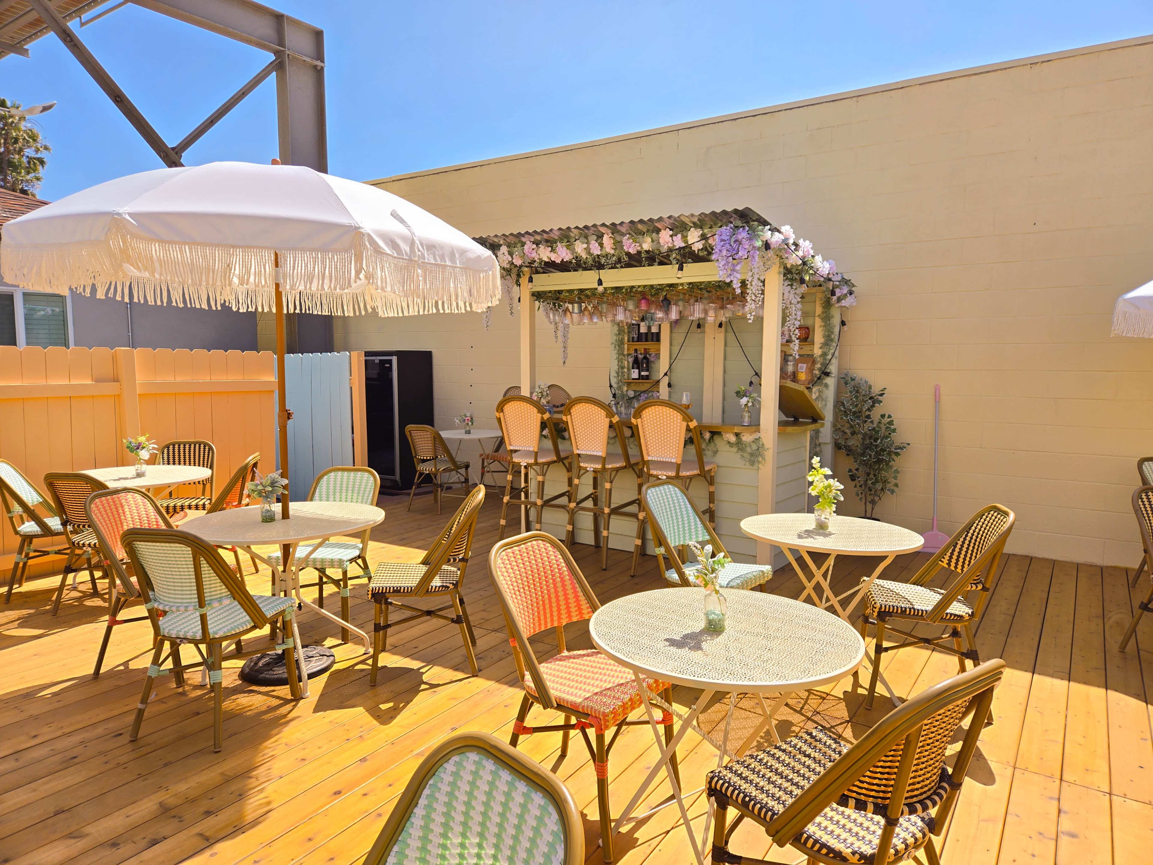 A sunny outdoor patio features colorful bistro tables and chairs arranged around a small bar decorated with flowers.