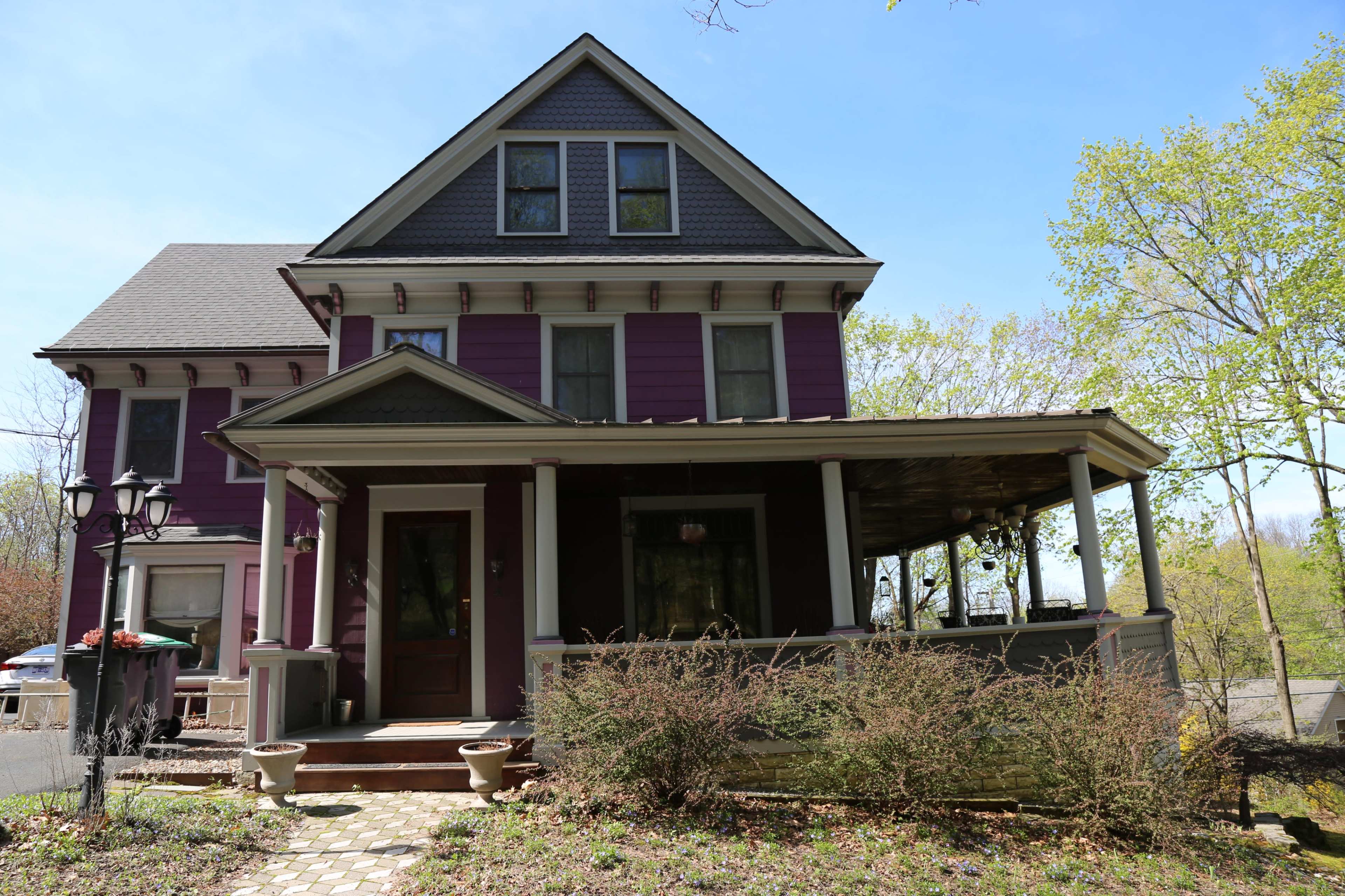 A large purple Victorian-style house with a front porch and a landscaped yard is set against a clear blue sky.