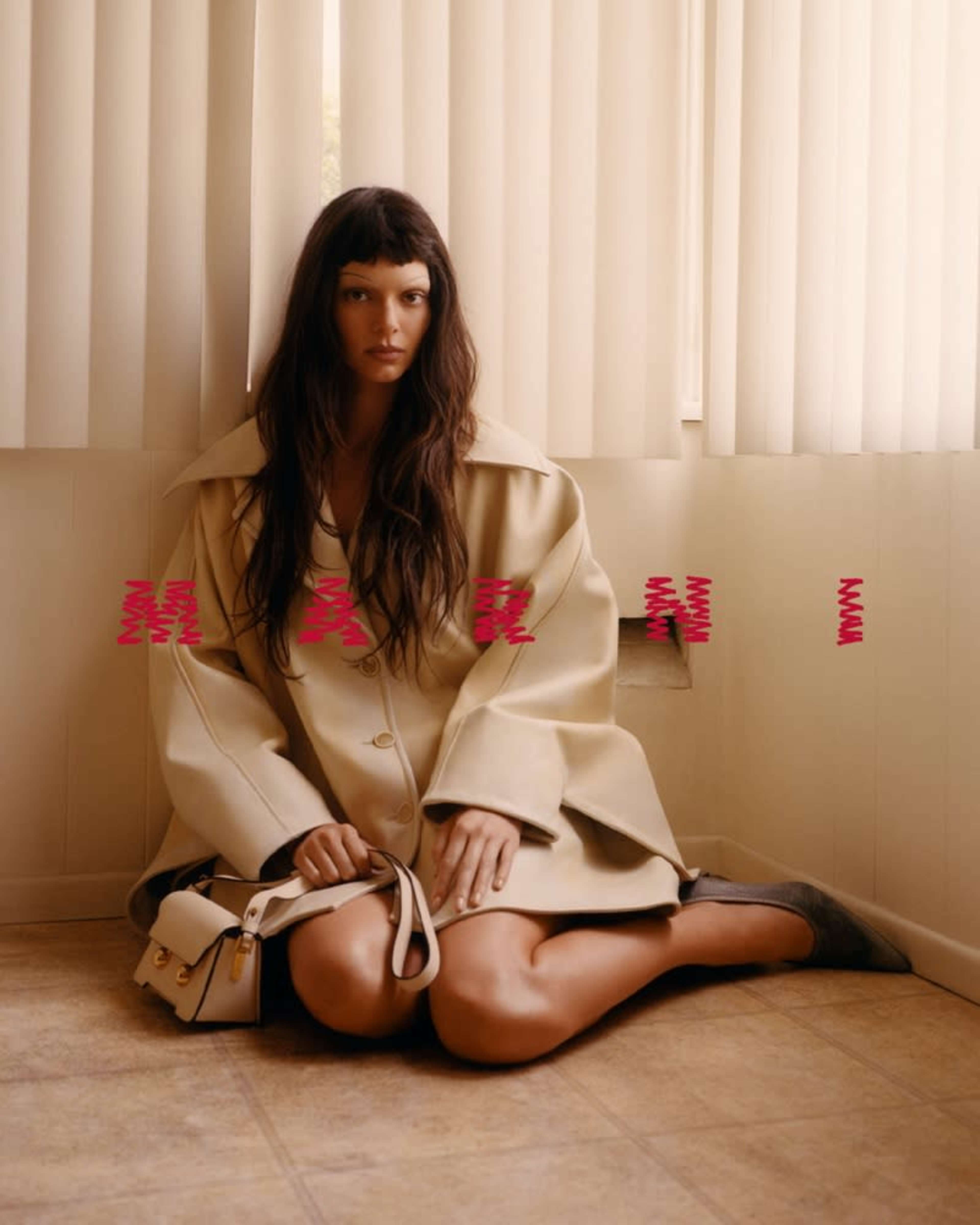 A woman sits on the floor in a beige coat against a wall with vertical blinds, holding a small handbag.