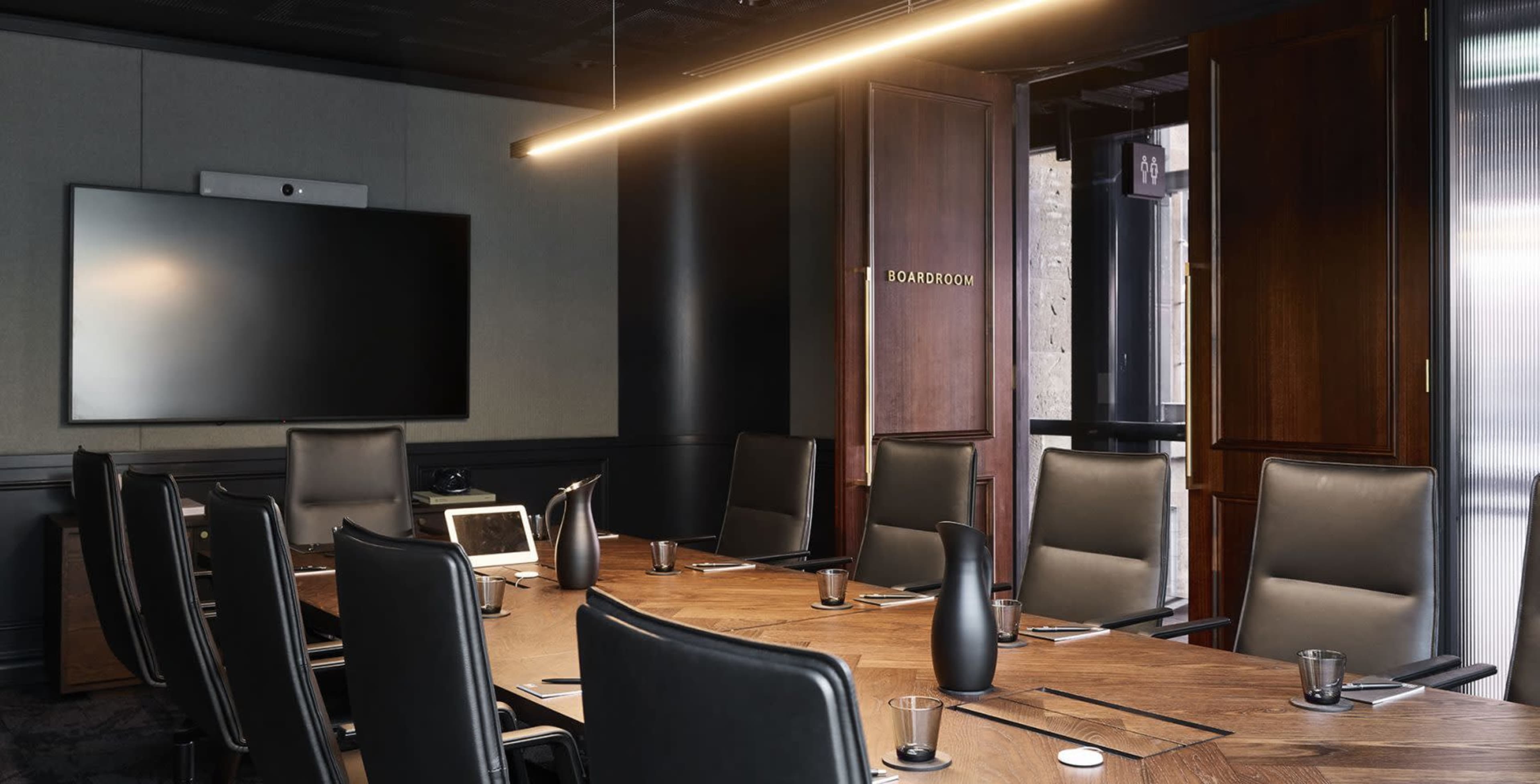 A modern boardroom with a long wooden table, black chairs, a large screen on the wall, and a door labeled "BOARDROOM."