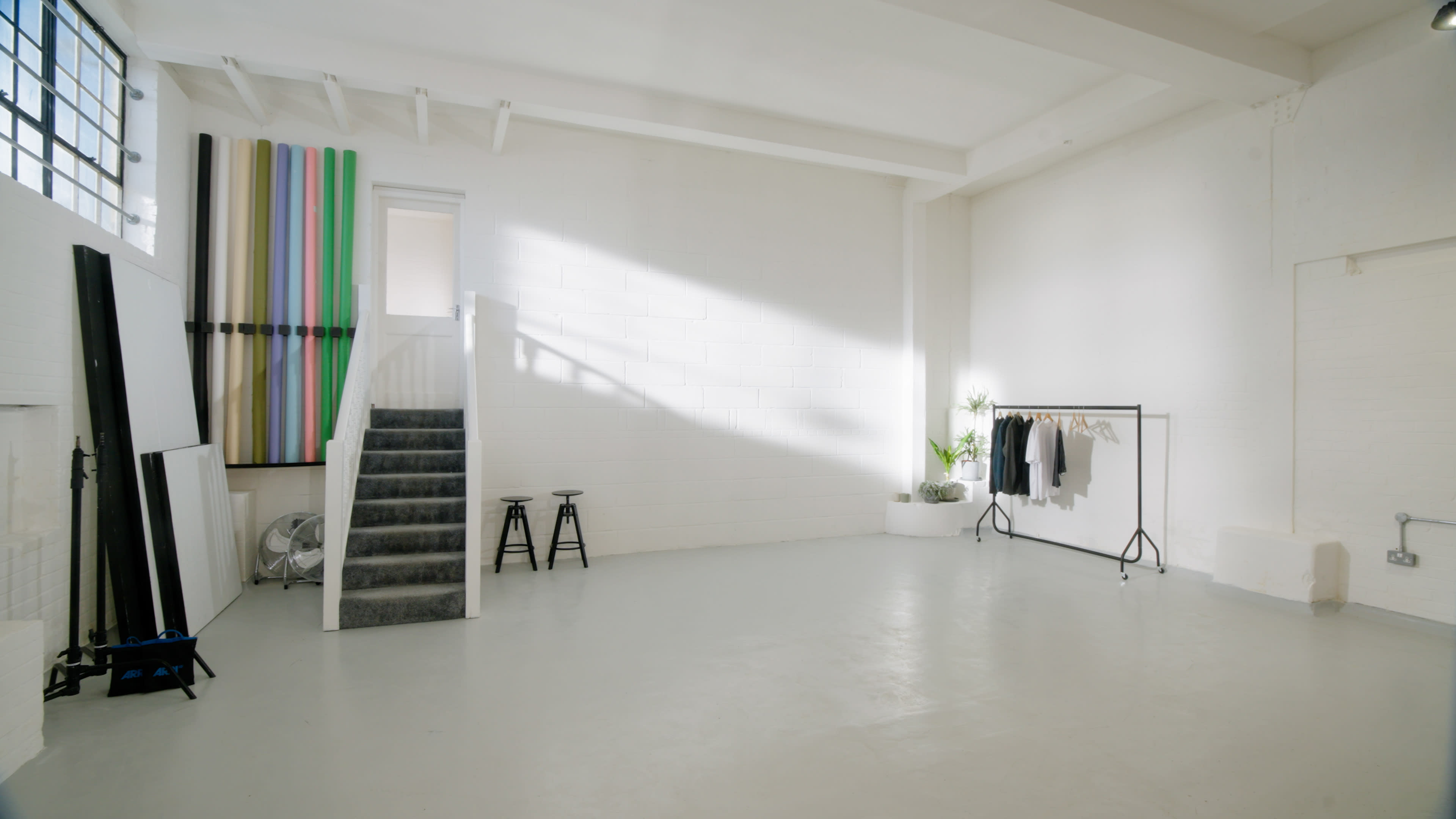 The image shows a bright, empty studio space with a staircase on one side and a clothing rack on the other.