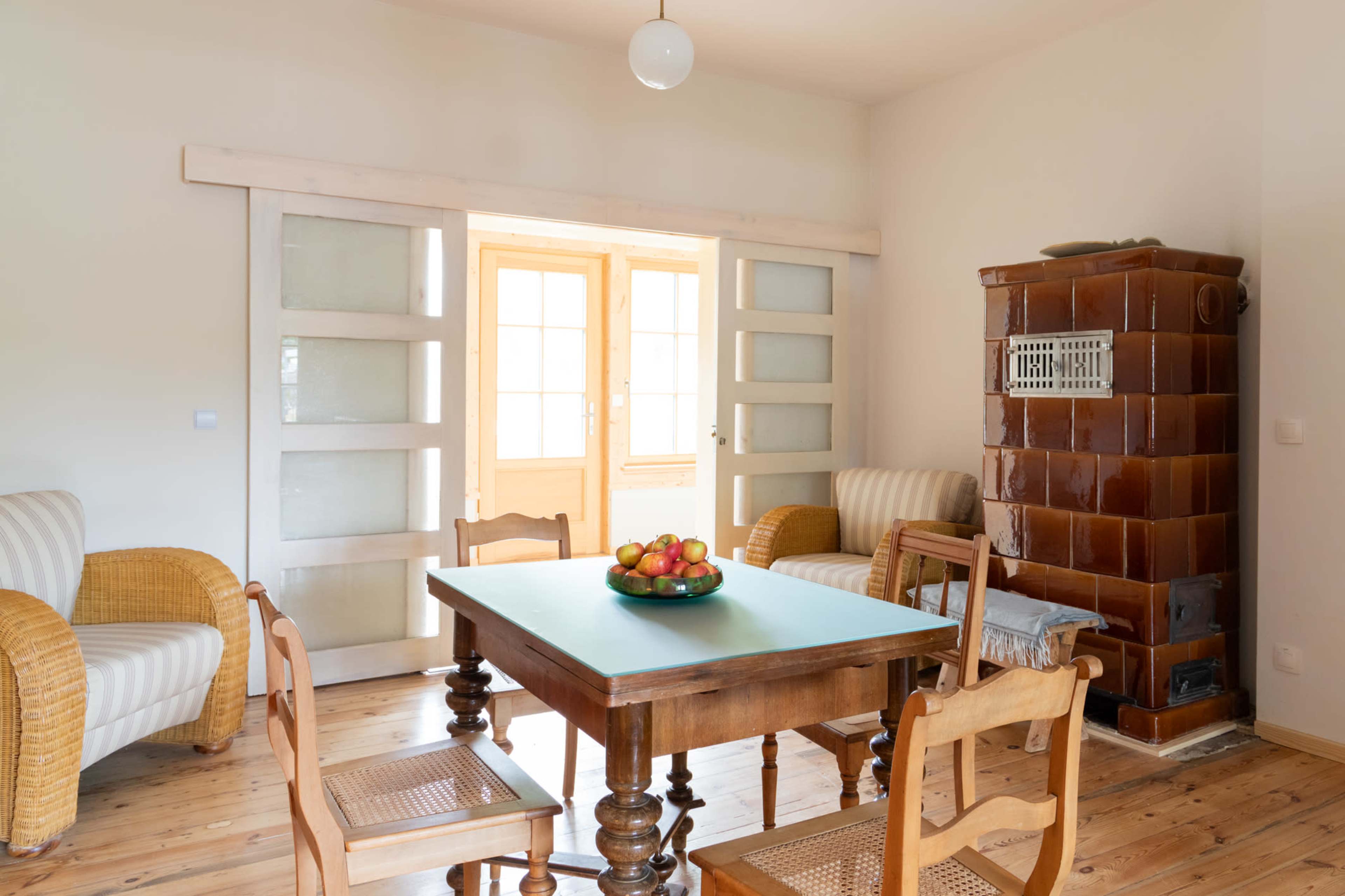 A simple dining area features a wooden table surrounded by chairs, a bowl of apples, and a tiled stove, with light streaming in through the open doors.