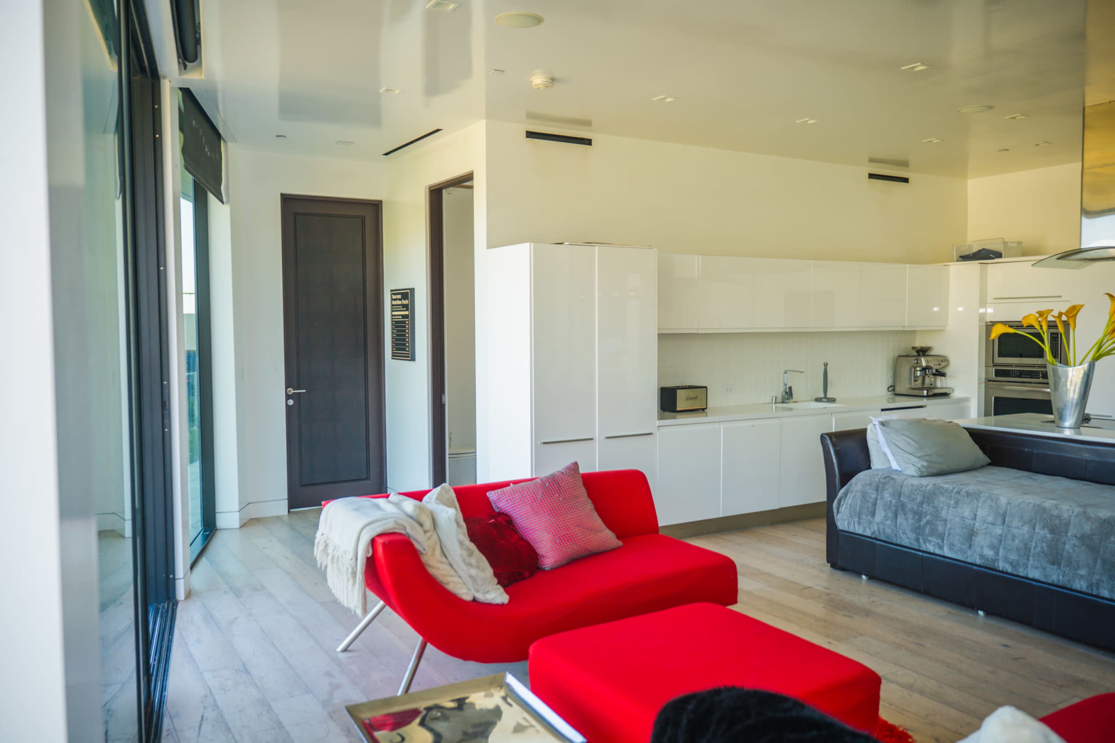 A modern living space features a red couch, a gray sectional, and a kitchen area with white cabinets and appliances.