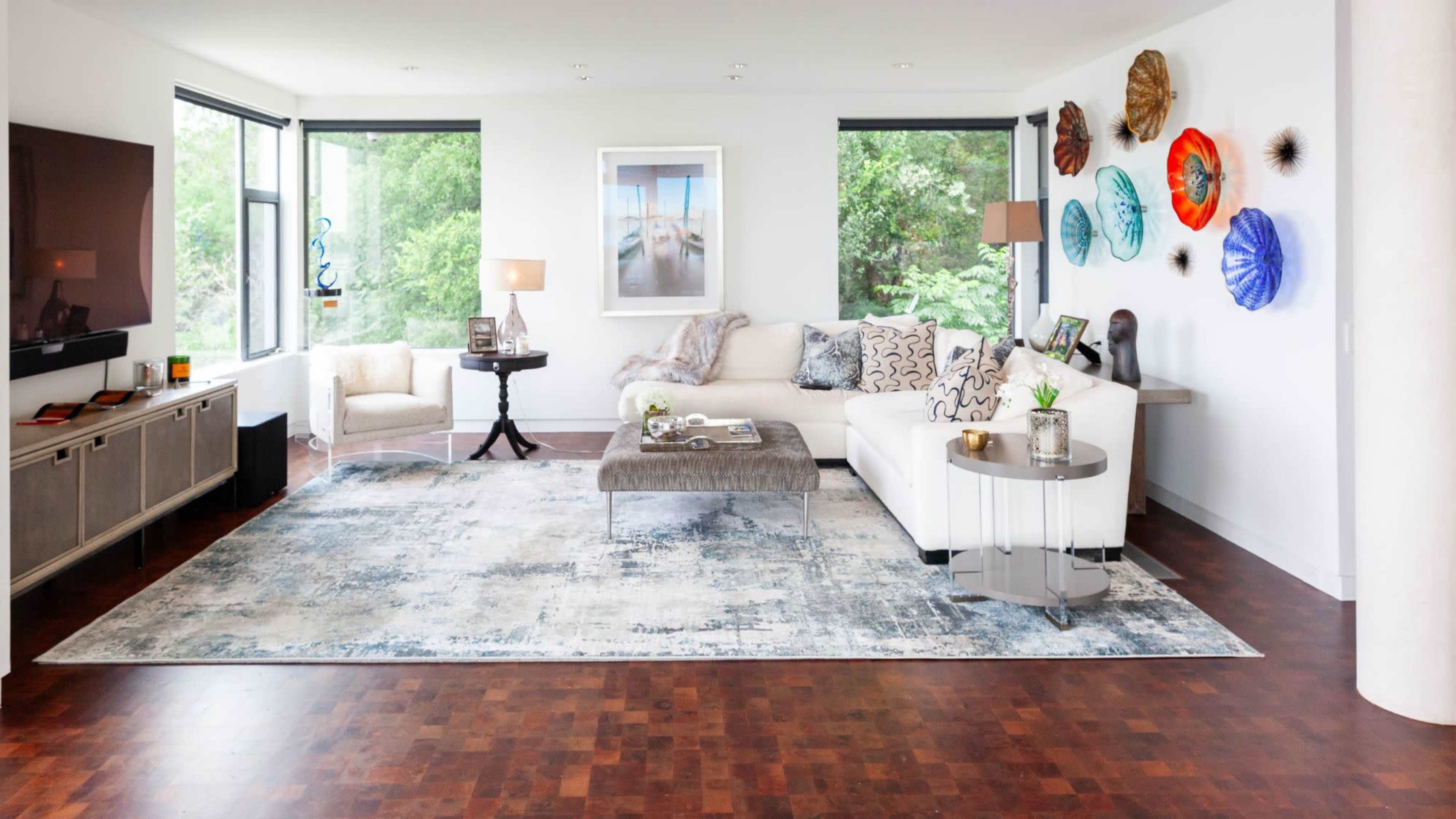 A modern living room features a white sofa, a gray coffee table, and decorative wall art, with large windows allowing natural light and views of greenery.