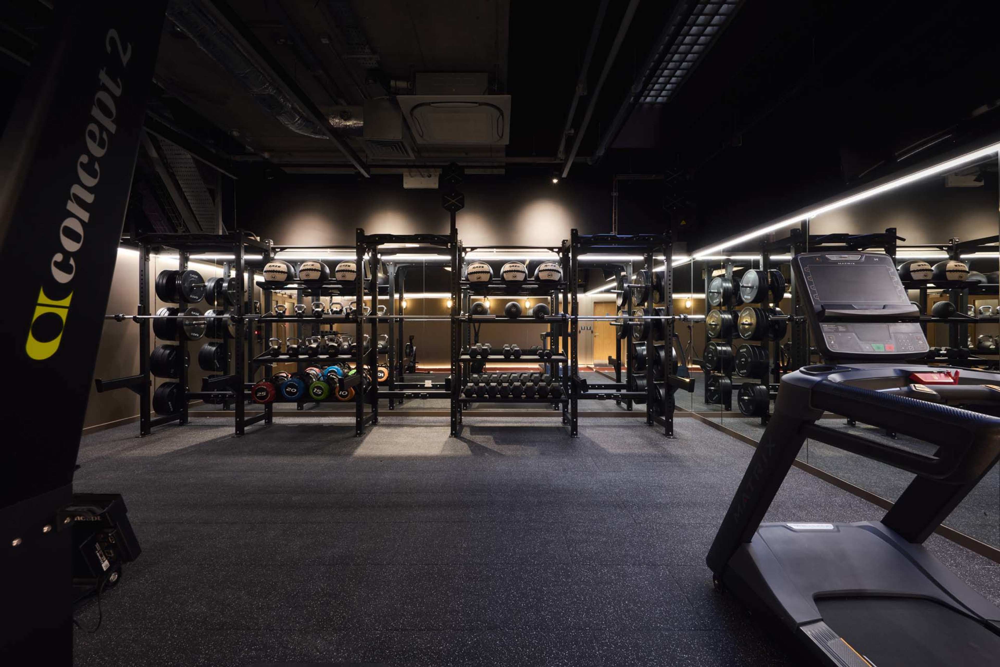 The image shows a modern gym interior featuring a treadmill on the right and a weight rack with various weights in the background.
