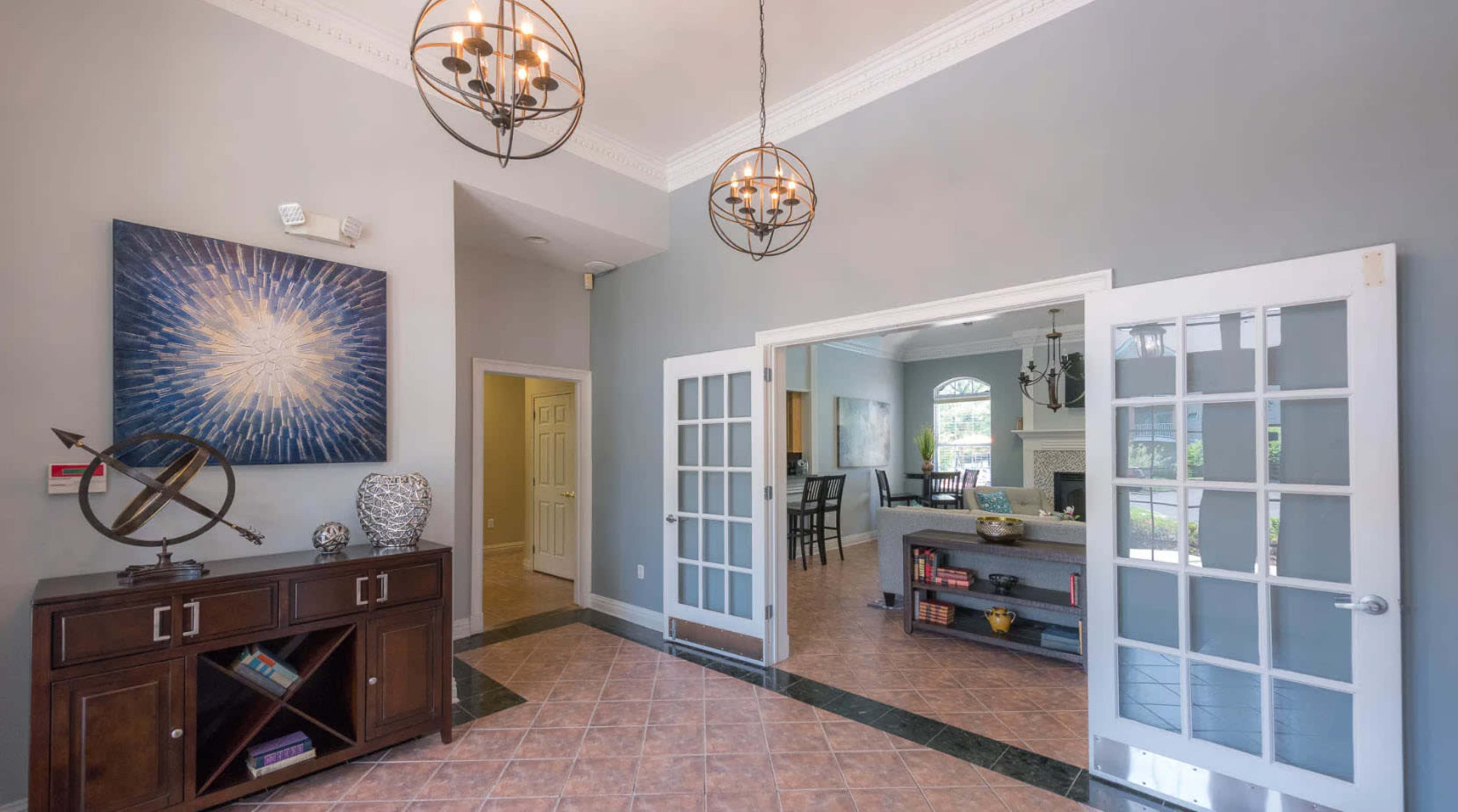 The image shows a spacious entryway with double doors leading to a bright living area, featuring a decorative wall art piece and a wooden cabinet on the left.