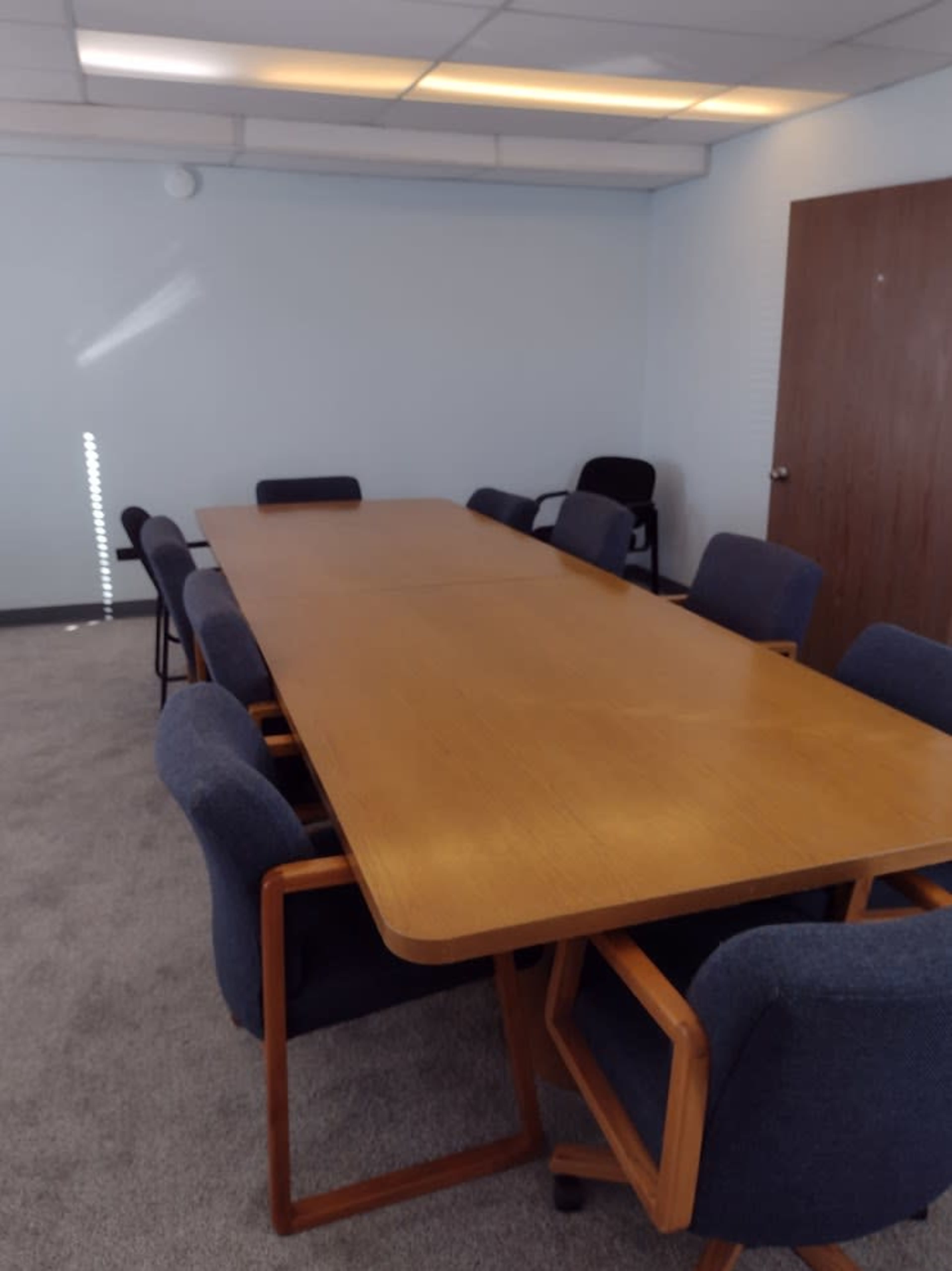 A long wooden conference table surrounded by upholstered chairs is set in a carpeted meeting room with a door and window.