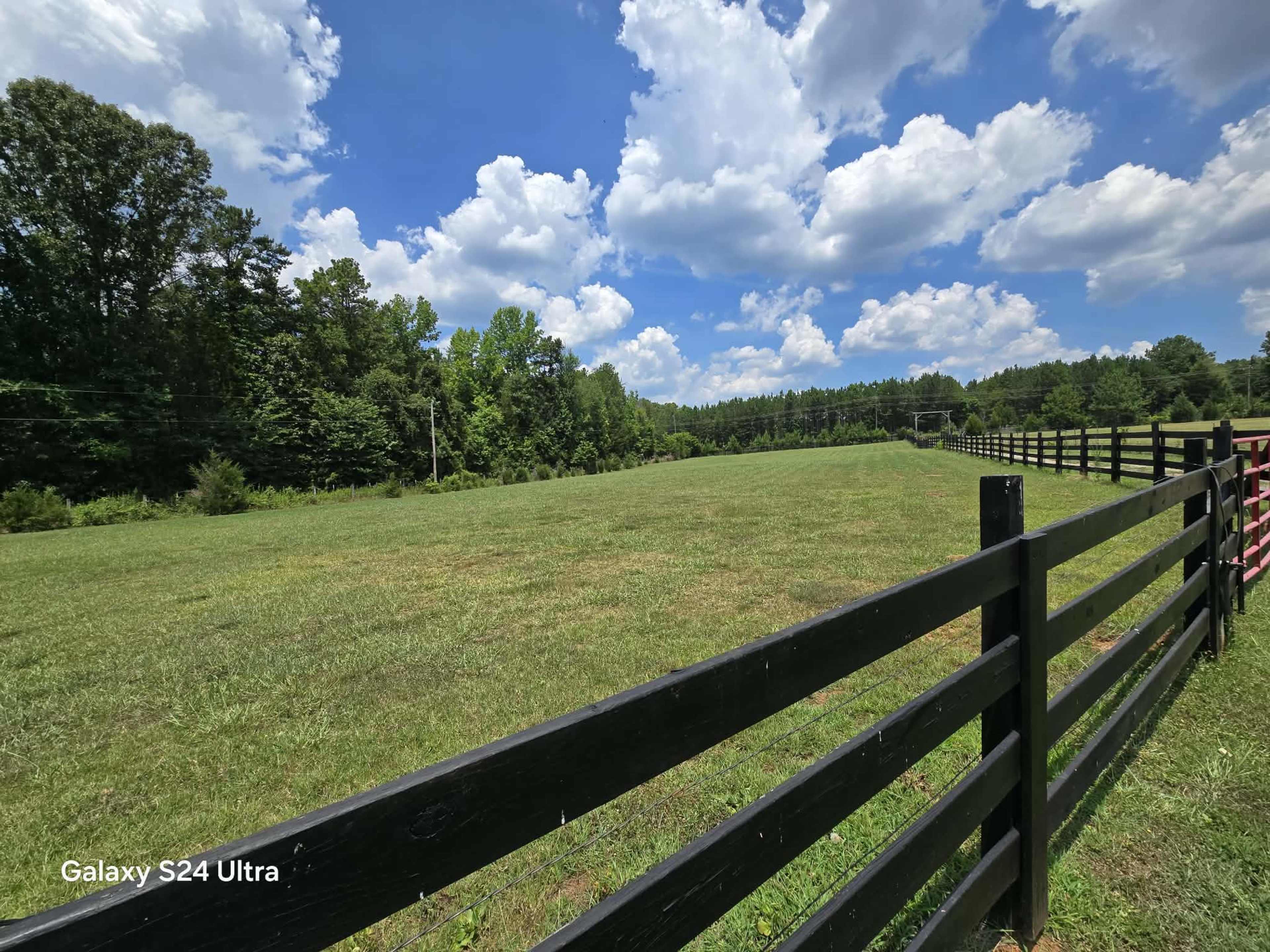 A black wooden fence lines a grassy field under a blue sky with scattered clouds.