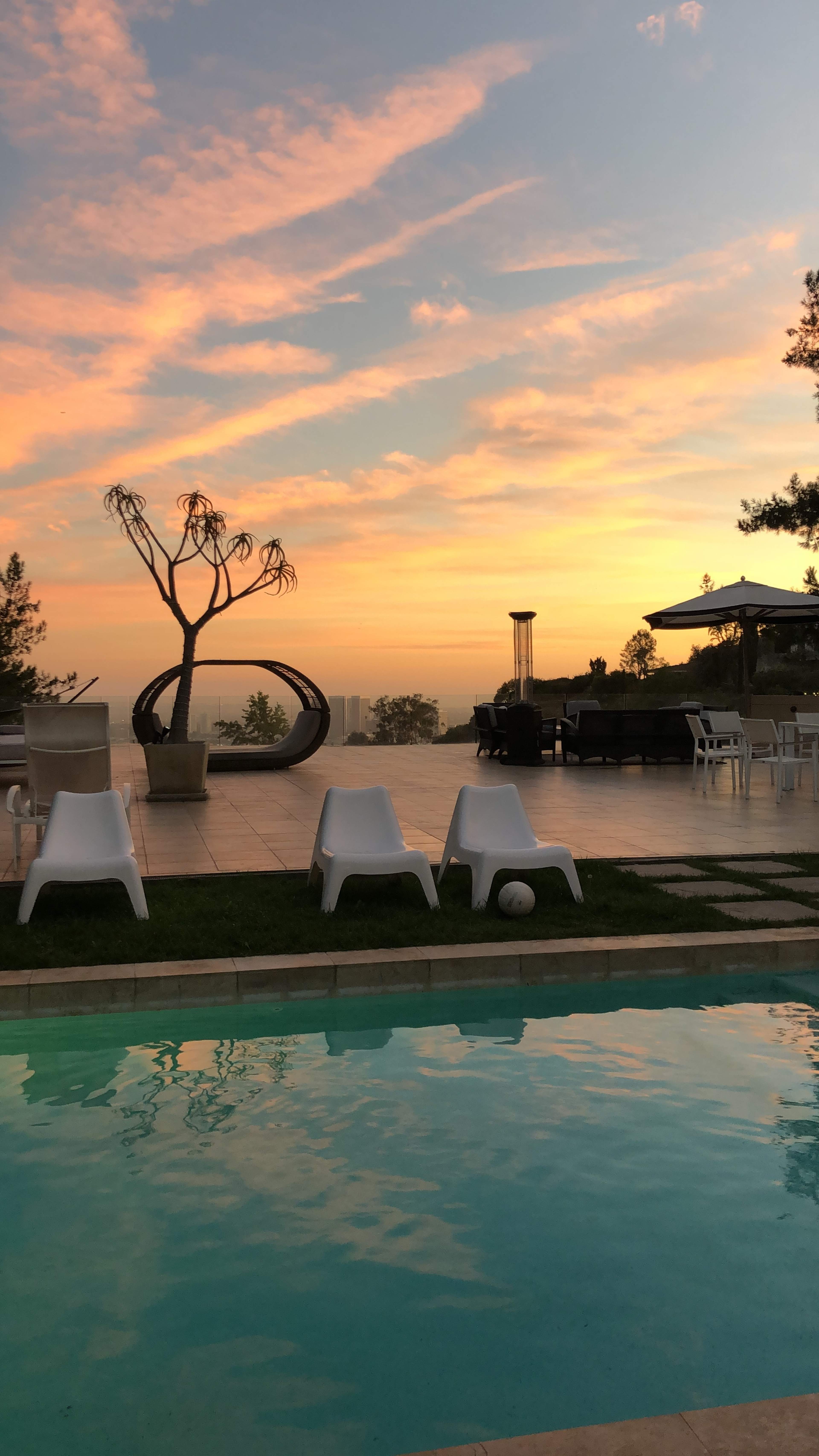 The image shows a poolside area with white chairs and a tree silhouetted against a sunset sky filled with clouds.