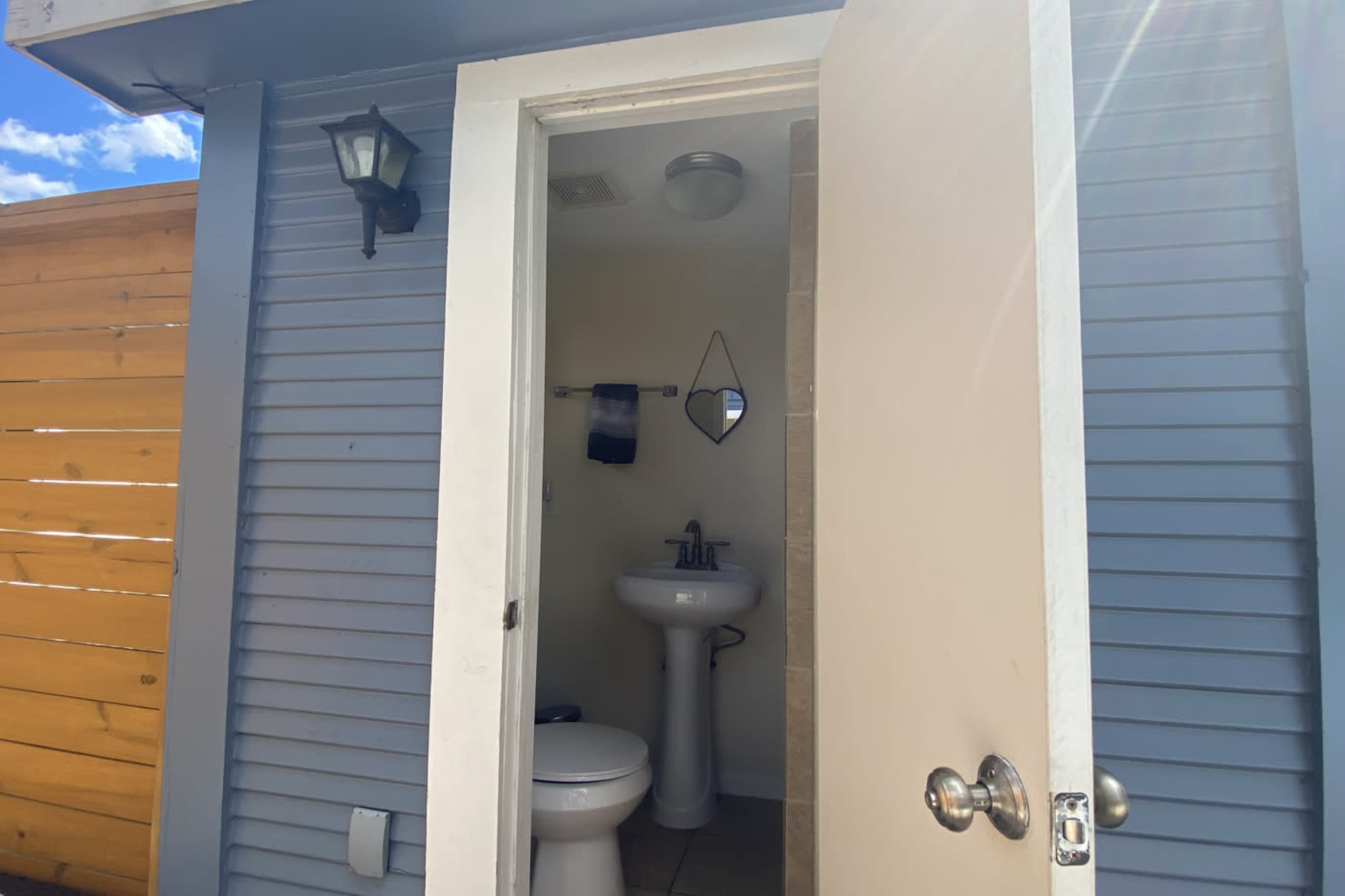 A bathroom is visible through an open door, featuring a sink, a toilet, and a mirror, with a wooden fence in the background.