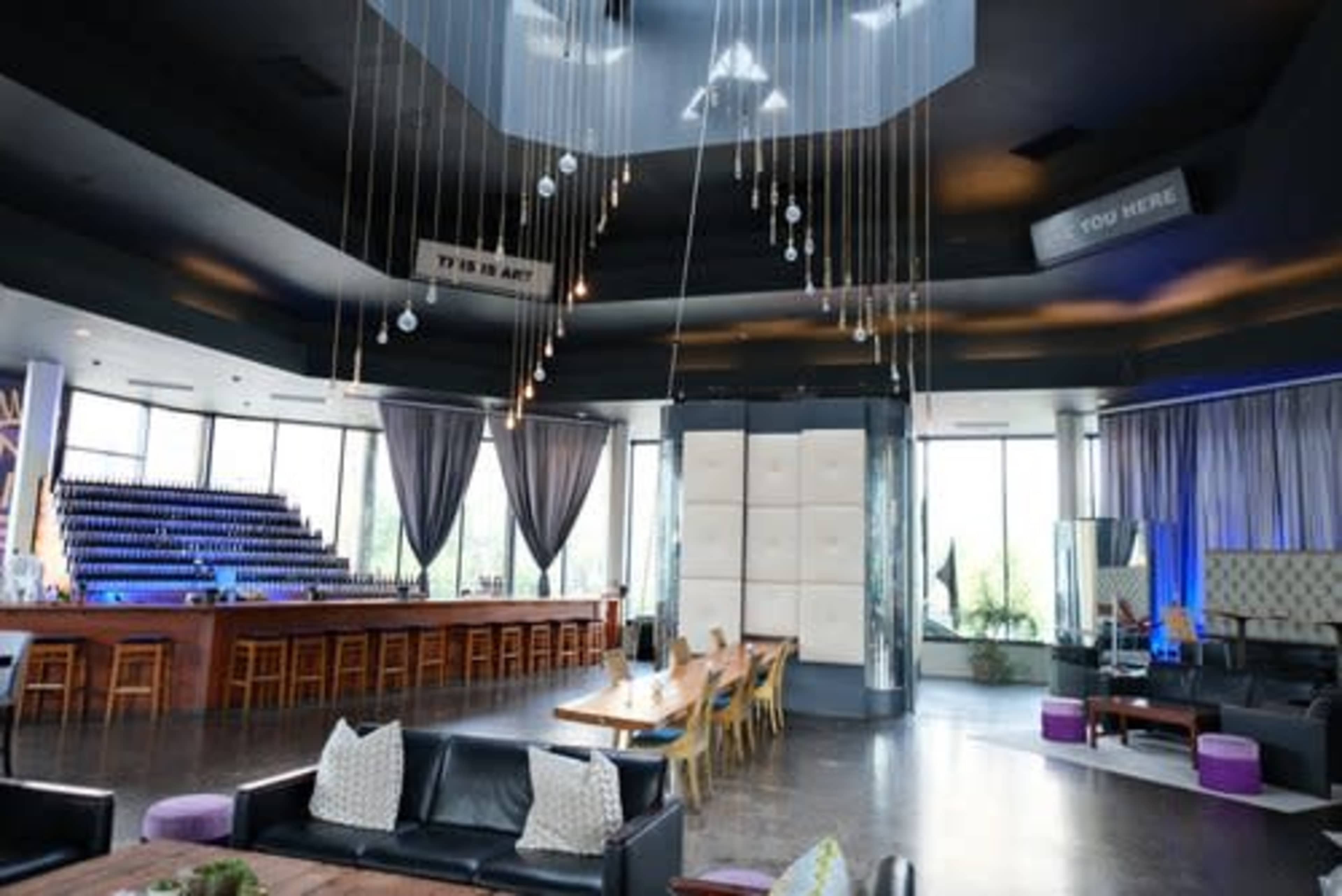 The image shows a modern interior of a bar or lounge with a sleek design, featuring large windows, hanging light fixtures, and a mix of seating arrangements.