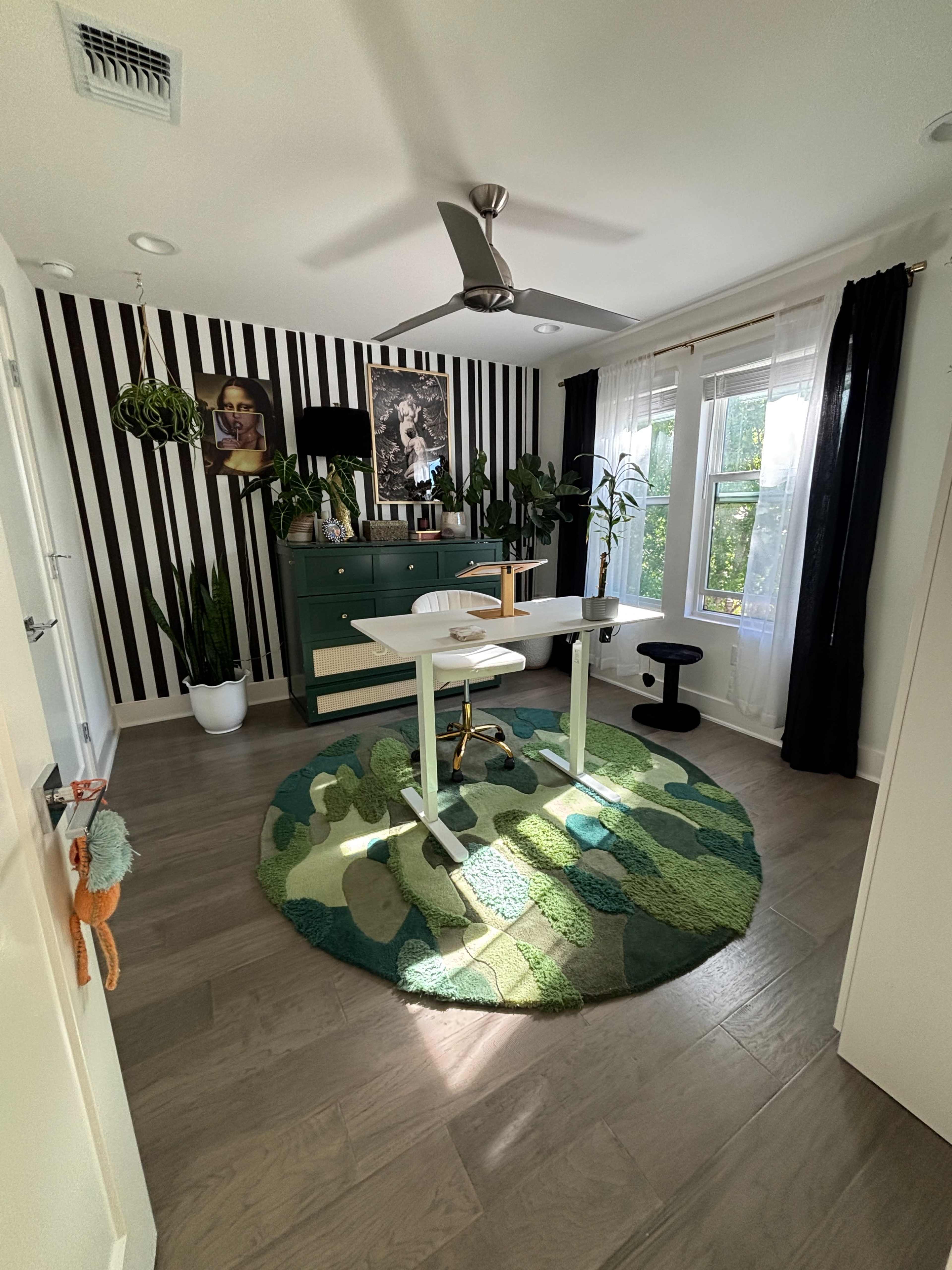 A brightly lit home office features a green chest of drawers, a round rug with a leaf pattern, plants, and a standing desk positioned near a window with white curtains.
