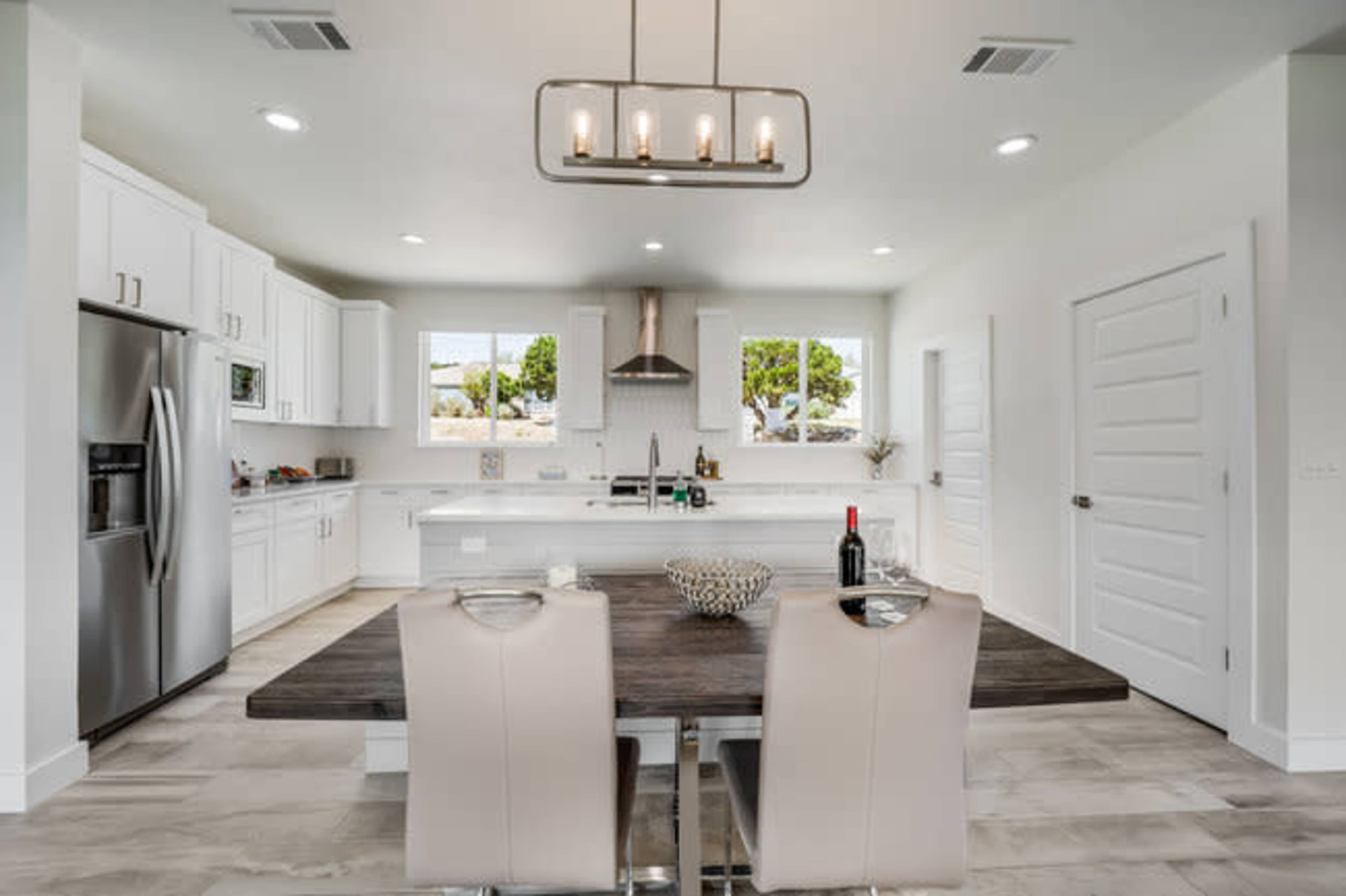 The image shows a modern kitchen and dining area featuring white cabinetry, stainless steel appliances, a large wooden dining table, and well-lit windows.